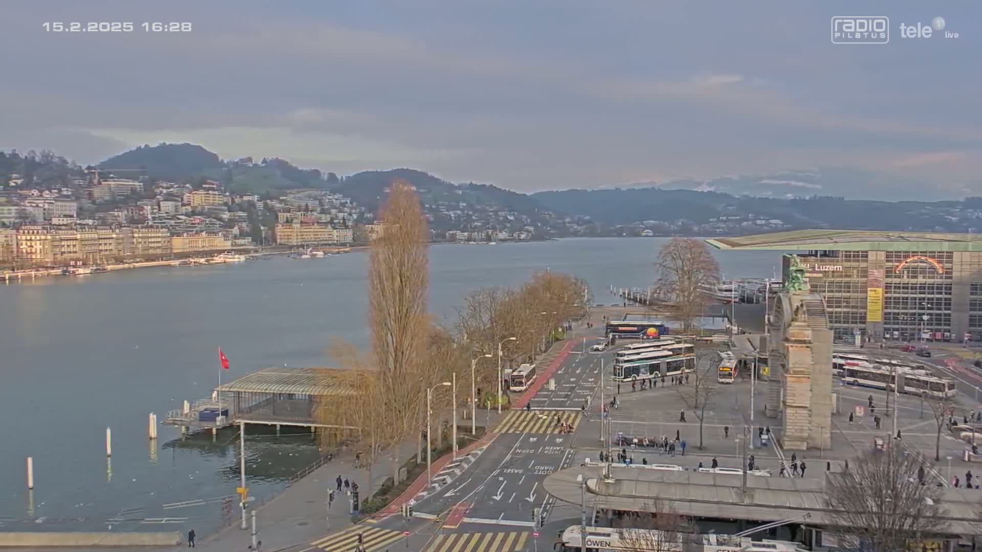 A city waterfront on an overcast day, with buses and pedestrians near a large building by the lake, and hills visible in the distance.
