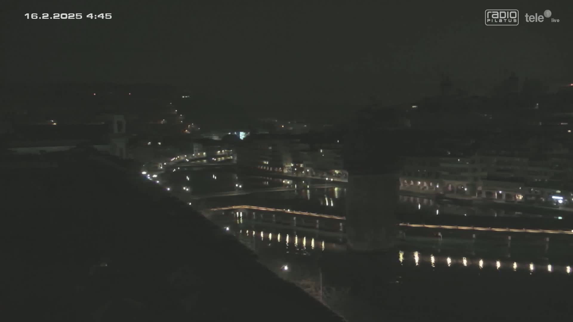 A nighttime, high-angle view shows a city beside a dark body of water, with lights illuminating buildings and the water's edge.