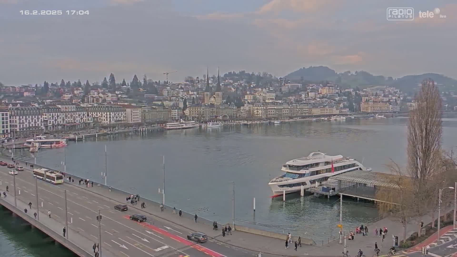 A city waterfront on an overcast day, showing a large passenger boat docked near a bridge with people walking along it and cars driving across.