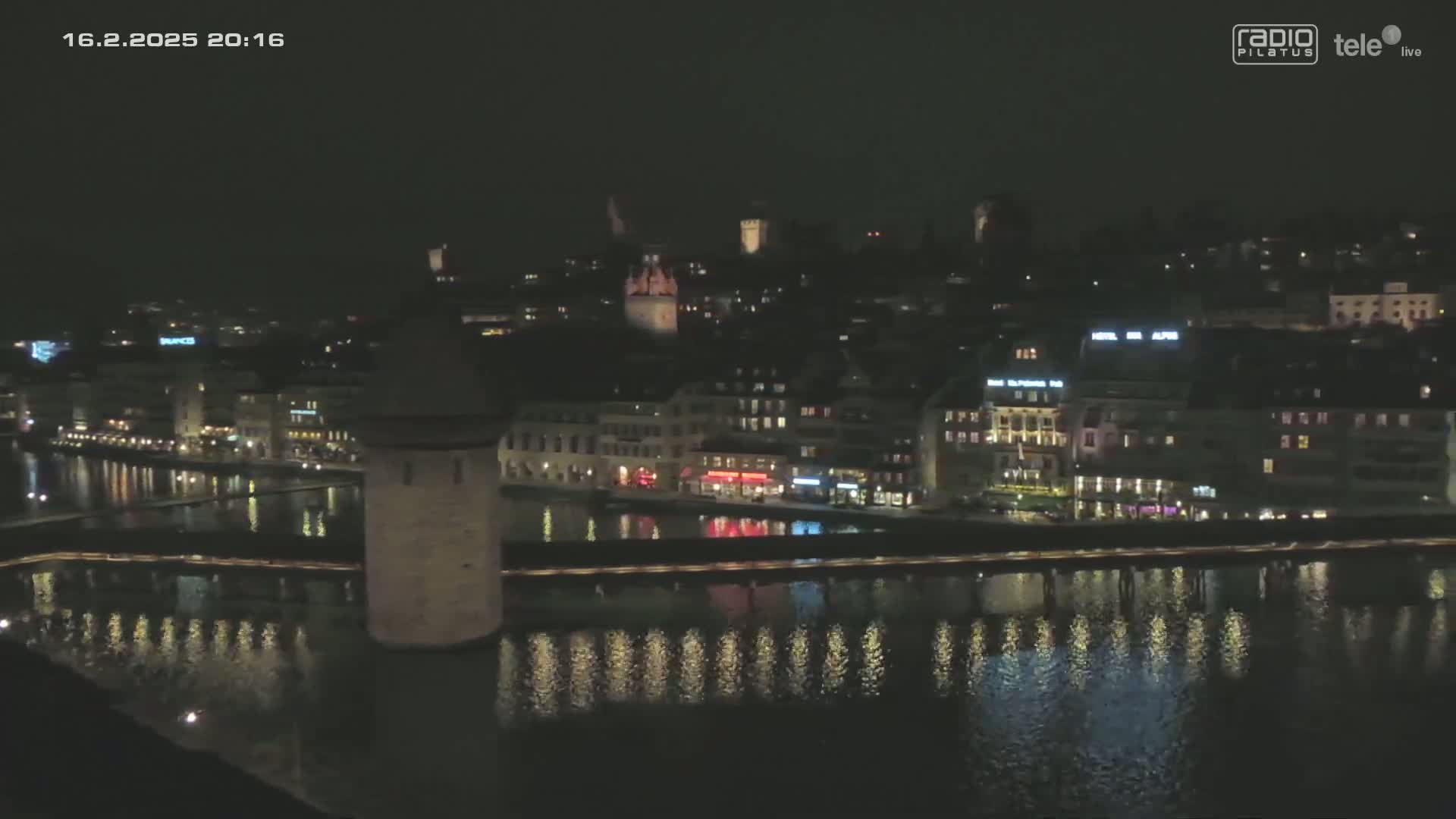 A nighttime view of a city's waterfront, showing buildings illuminated along a river with their lights reflecting on the calm water, a stone tower near the riverbank, and a bridge connecting to the opposite shore.