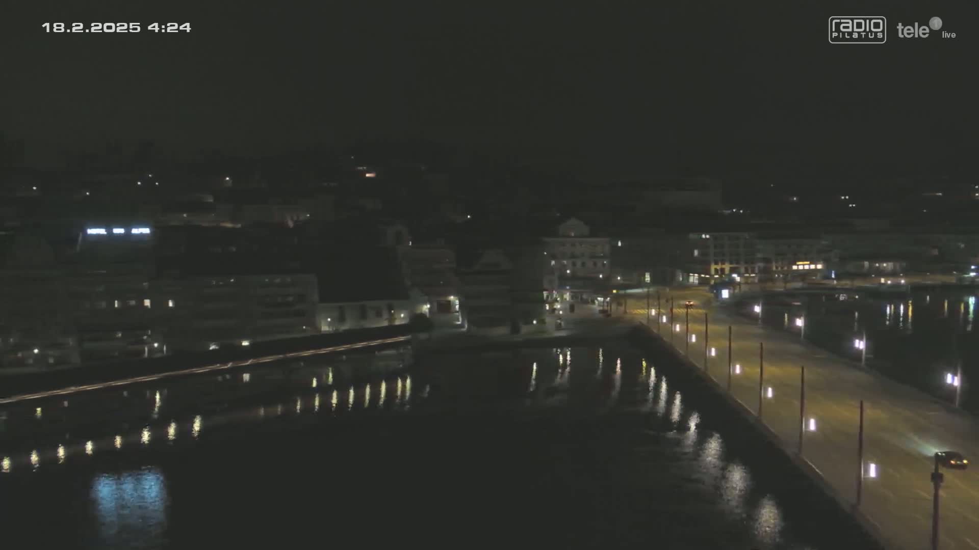 A nighttime, high-angle view shows a city waterfront with a bridge illuminated by streetlights, reflecting in calm water.