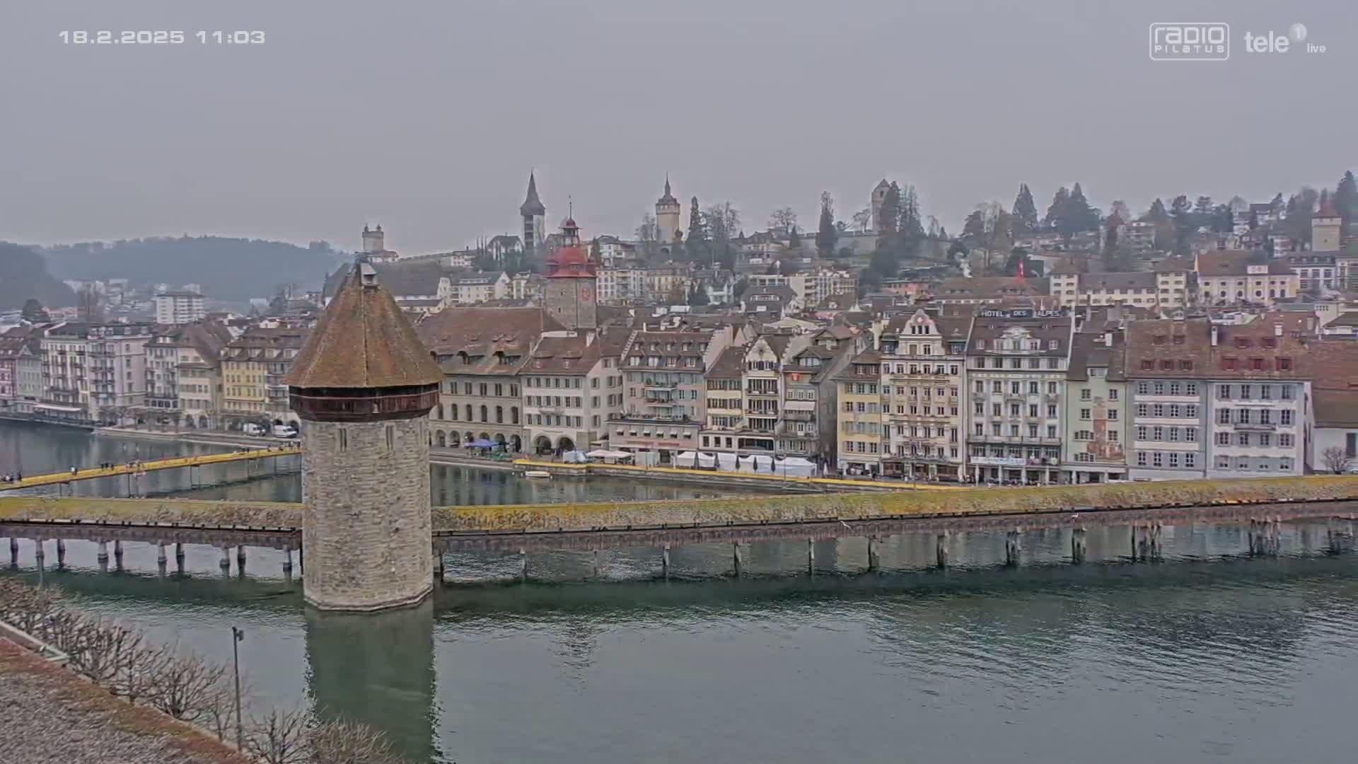 A stone water tower sits on a bridge over a calm river in front of a city of closely packed buildings under an overcast sky.