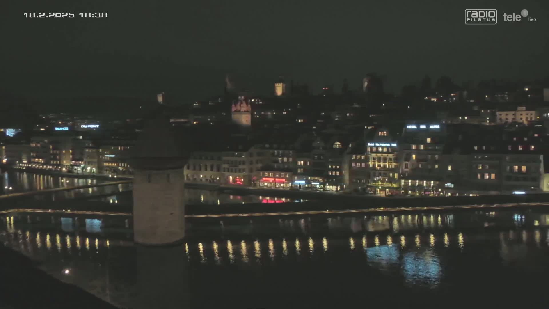 A nighttime view of a city's waterfront, featuring illuminated buildings and a stone tower reflecting in calm water.