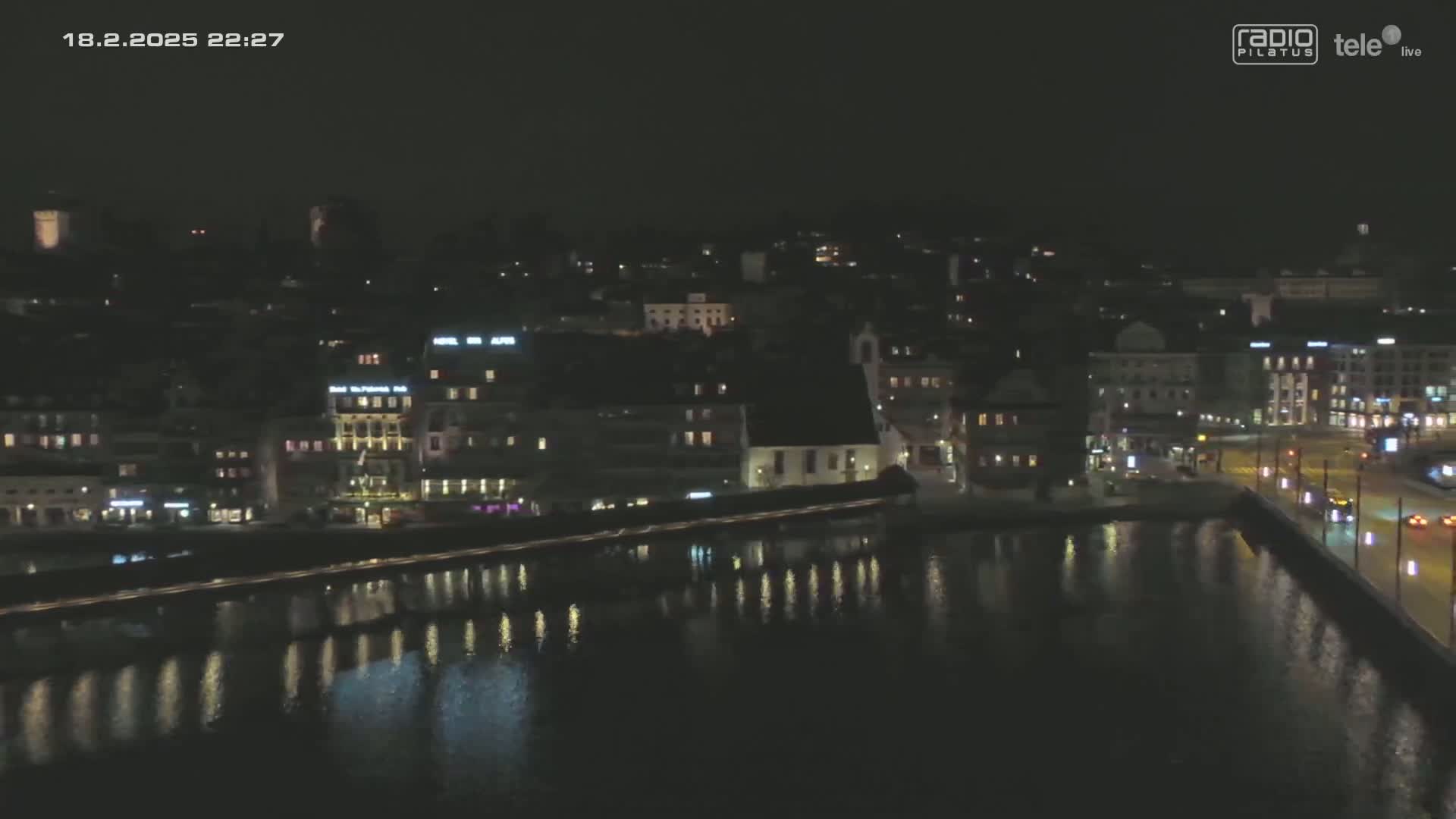 A nighttime, aerial view of a city waterfront, showing buildings with lights reflecting on calm water.