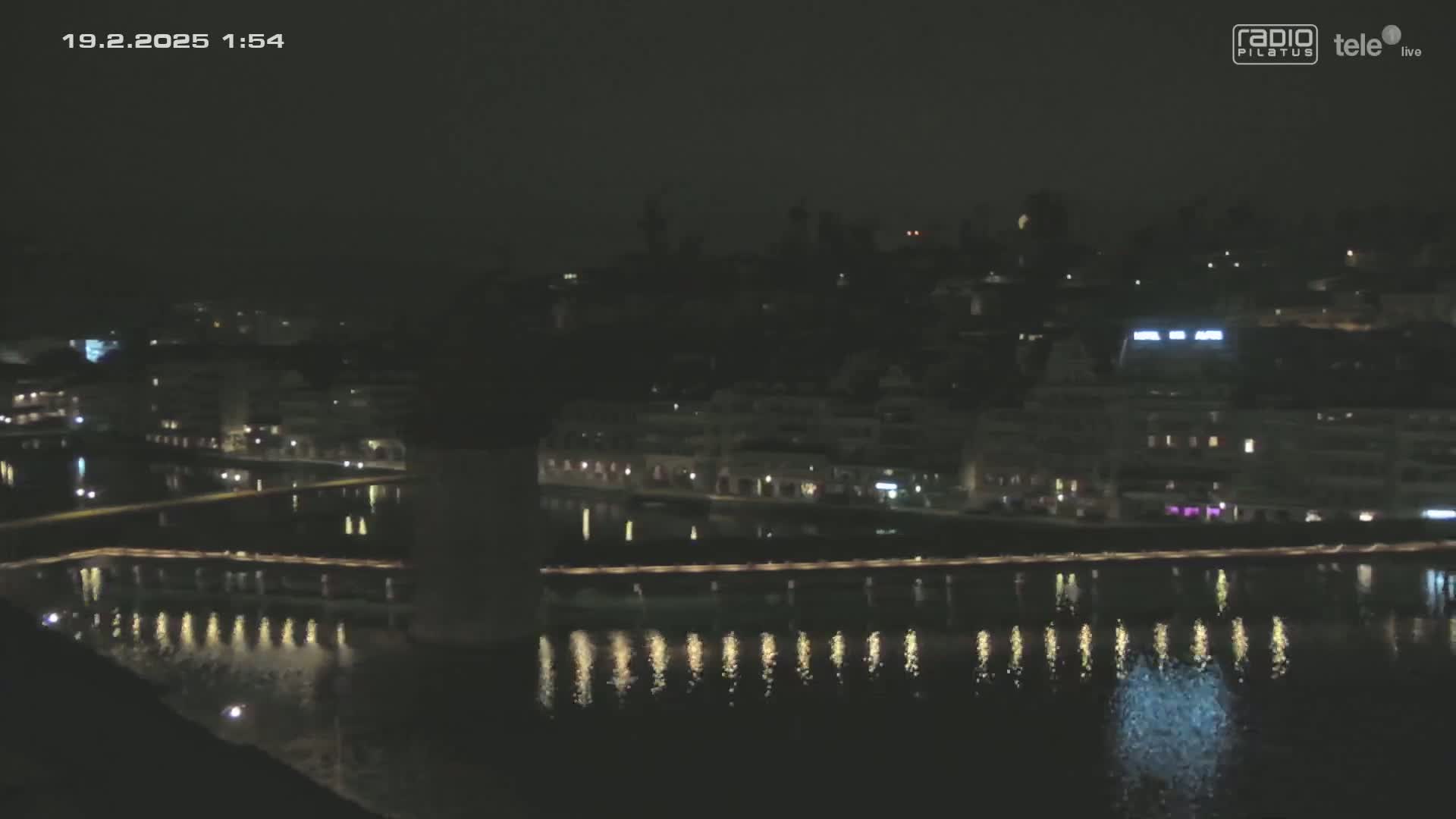 A nighttime, dark city skyline is reflected in a calm body of water, with lights illuminating the waterfront.