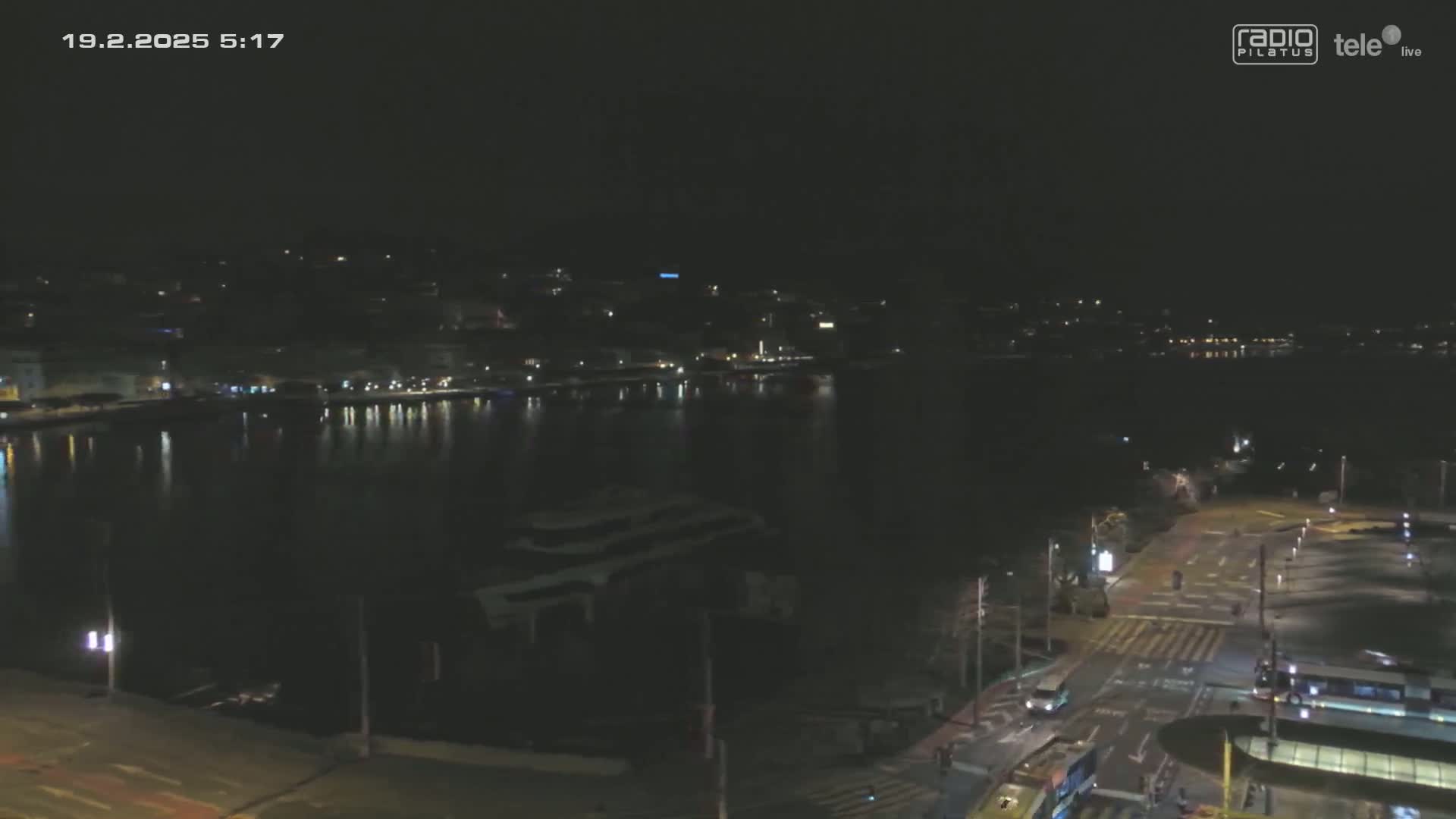 A nighttime, dark view of a calm body of water reflecting a city skyline, with a road and some vehicles visible in the foreground.
