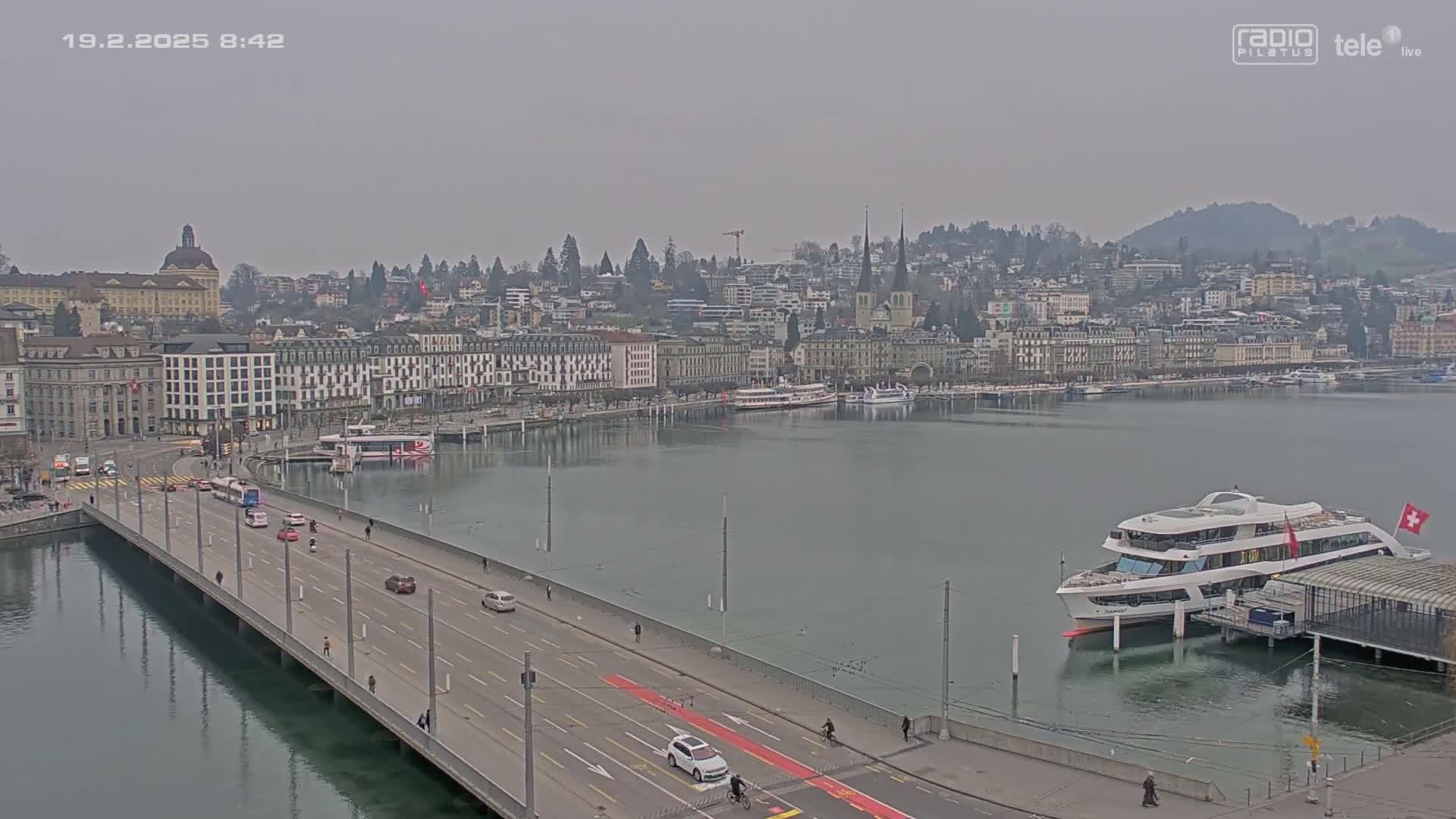 A bridge over calm water leads to a city with numerous buildings along a waterfront, under an overcast sky.