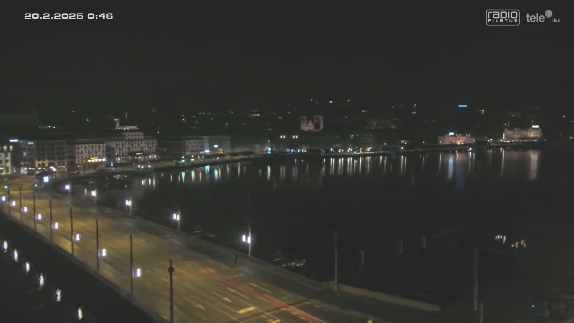 A nighttime view of a city's waterfront, showing a bridge with lights and buildings reflecting in calm water.