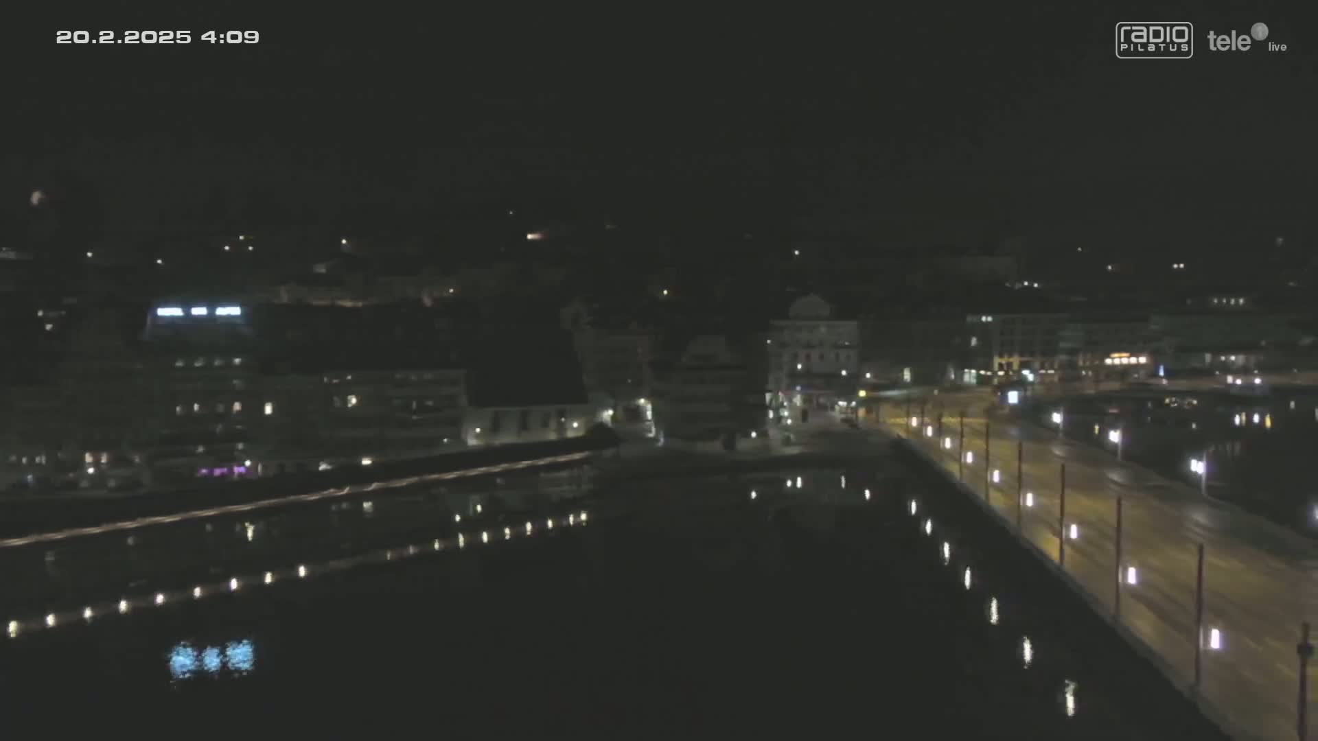 A nighttime, high-angle view shows a city bridge illuminated with streetlights, reflecting in dark water, alongside buildings with some lights on under a clear, dark sky.