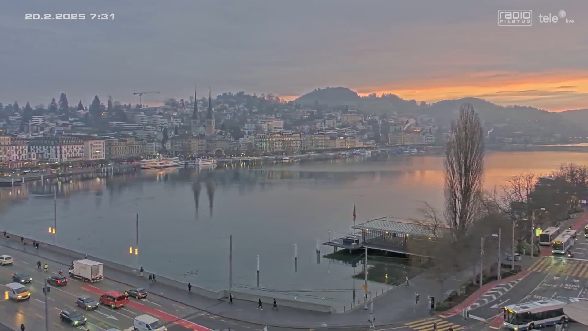 A city waterfront at sunrise, with calm water reflecting buildings and a partly cloudy sky, shows traffic on a roadway beside the water.