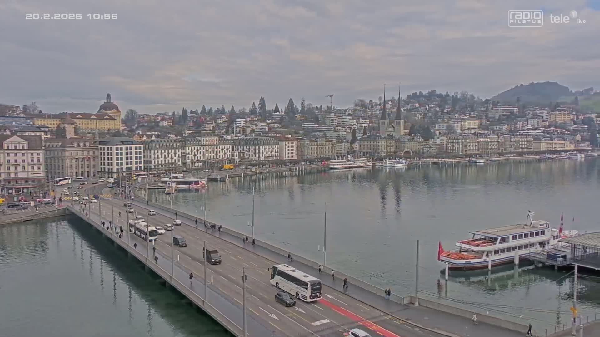 A bridge over calm water leads to a city of multi-story buildings under a cloudy sky.