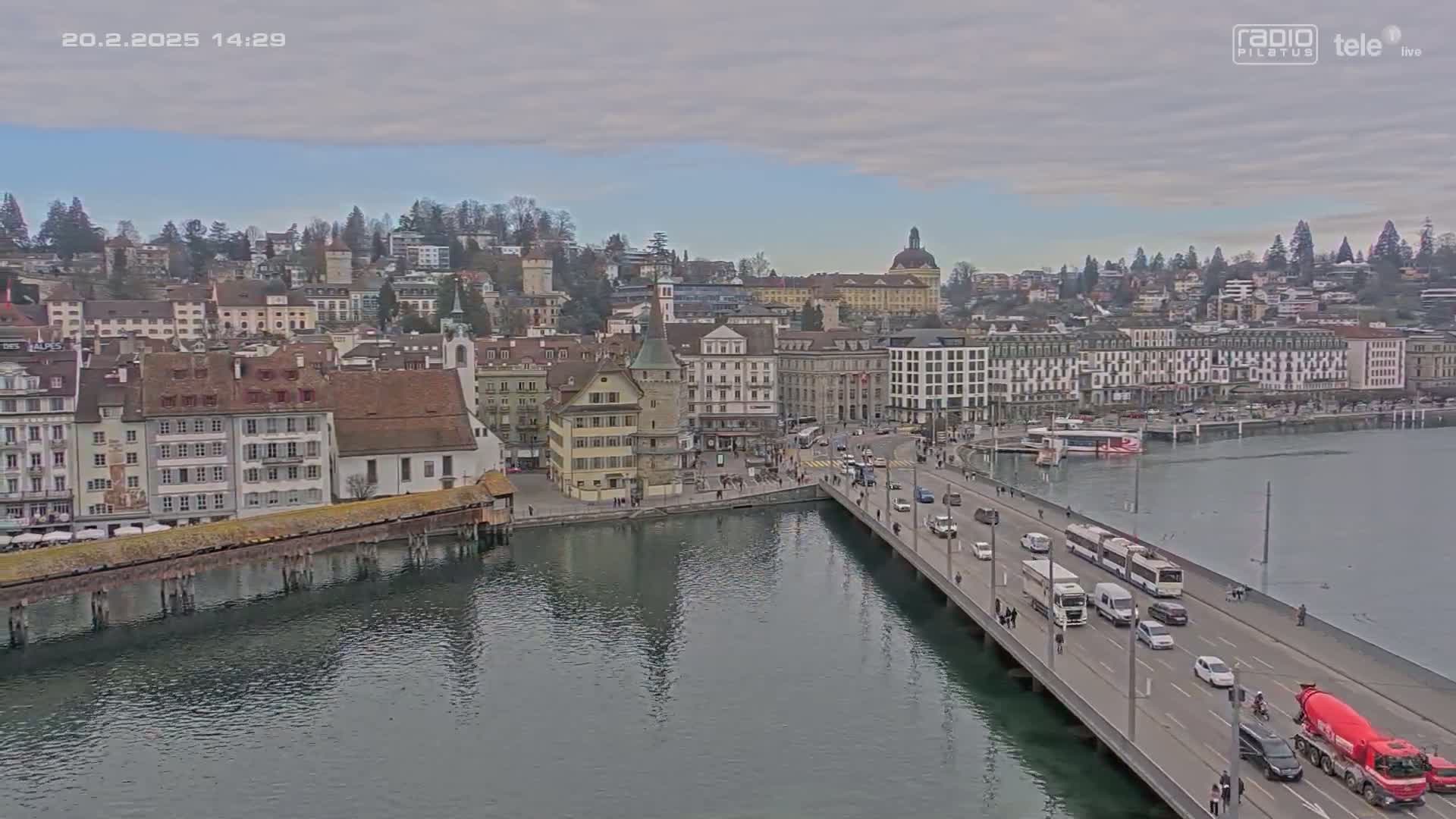 A bridge over a calm body of water leads to a town of European-style buildings under a partly cloudy sky.