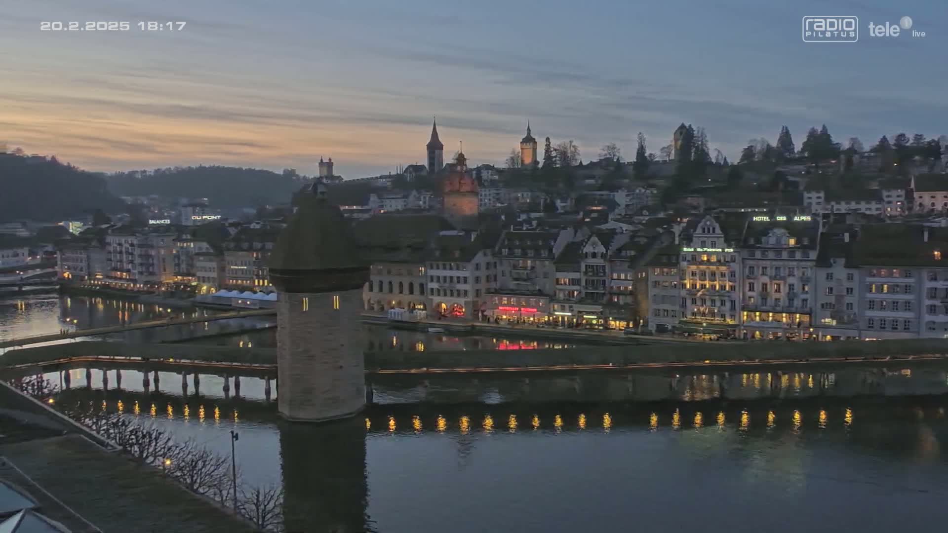 A twilight view of a European city with a river reflecting the lights of buildings along its banks, a stone tower on a bridge, and hills in the background.