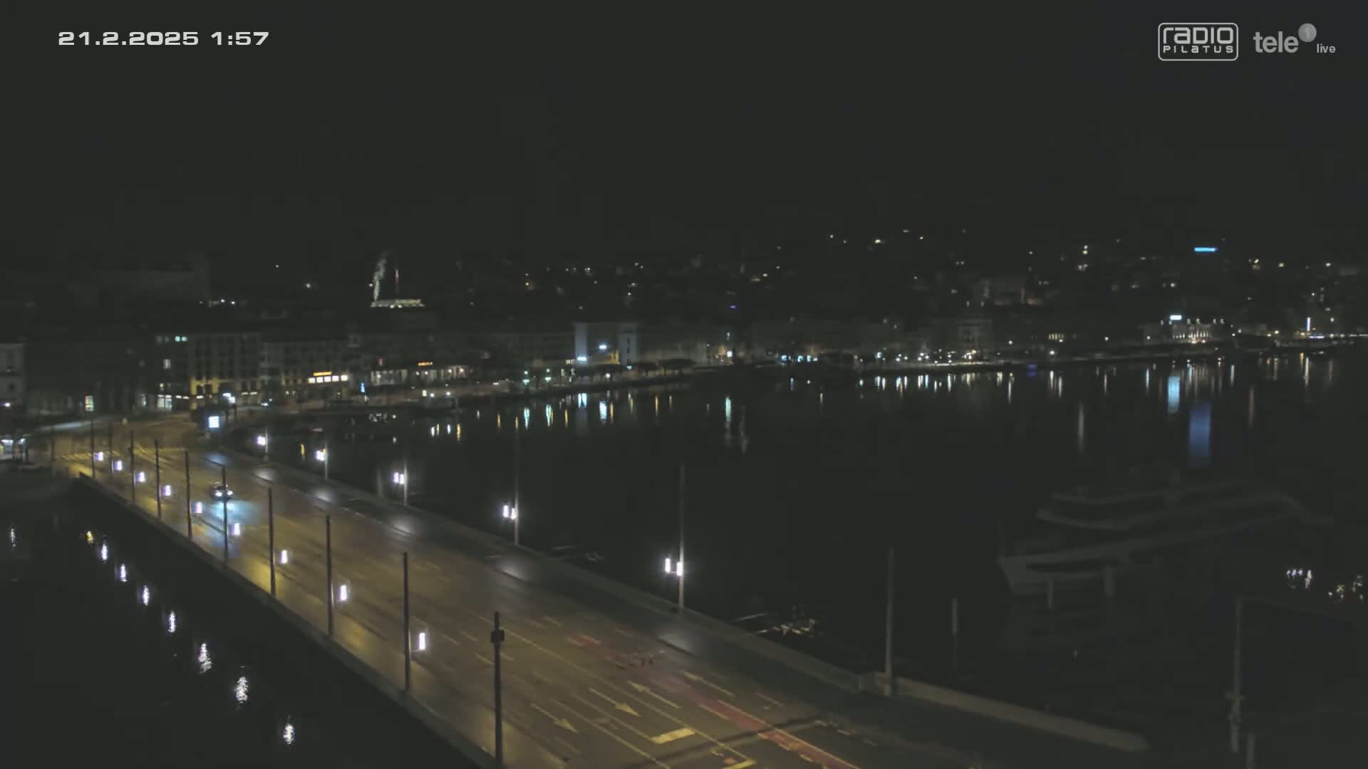 A nighttime view of a city's waterfront, showing a bridge with cars driving on it, buildings reflected in calm water, and a boat moored nearby.