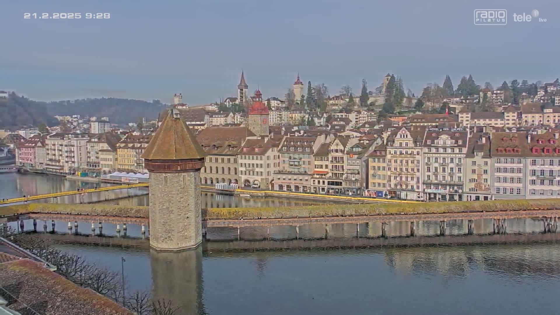 A stone water tower sits on a bridge spanning a calm river reflecting a cityscape of multi-story buildings under a clear sky.