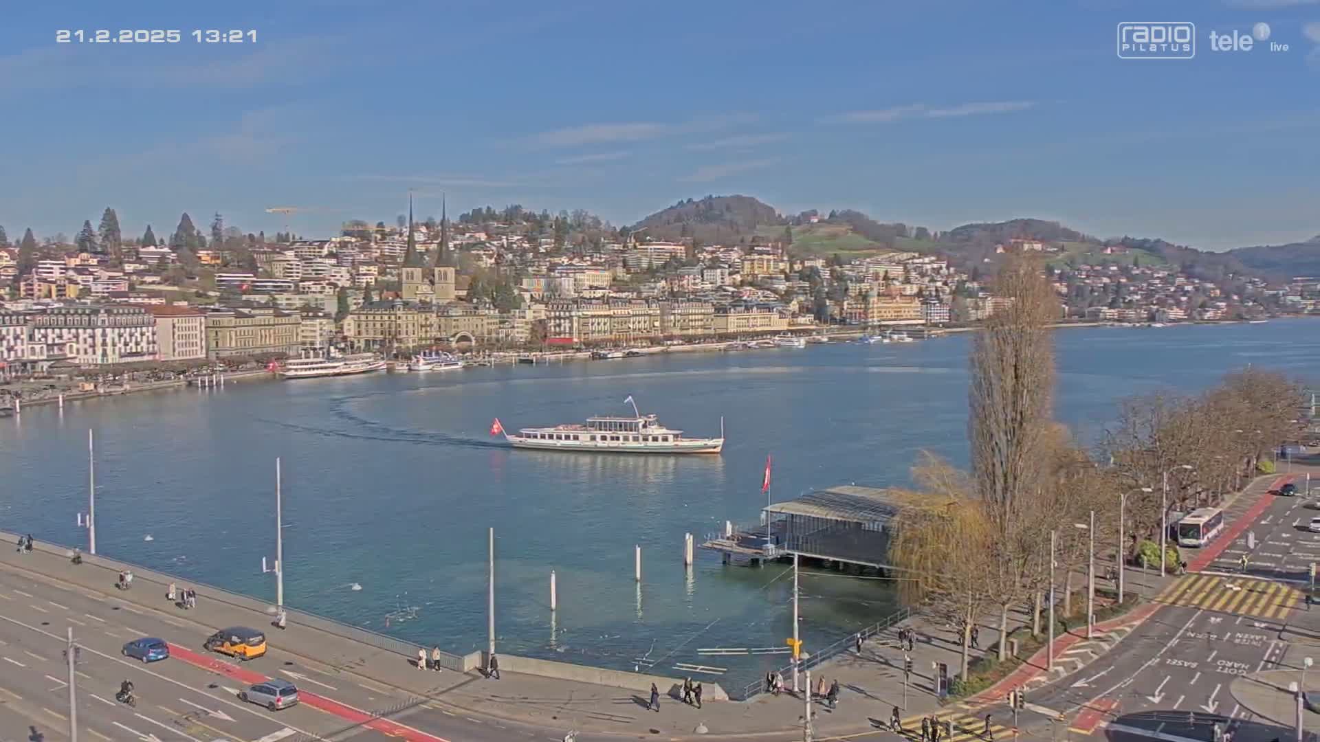 A large passenger boat travels across a calm lake in front of a town situated on a hillside under a clear blue sky.
