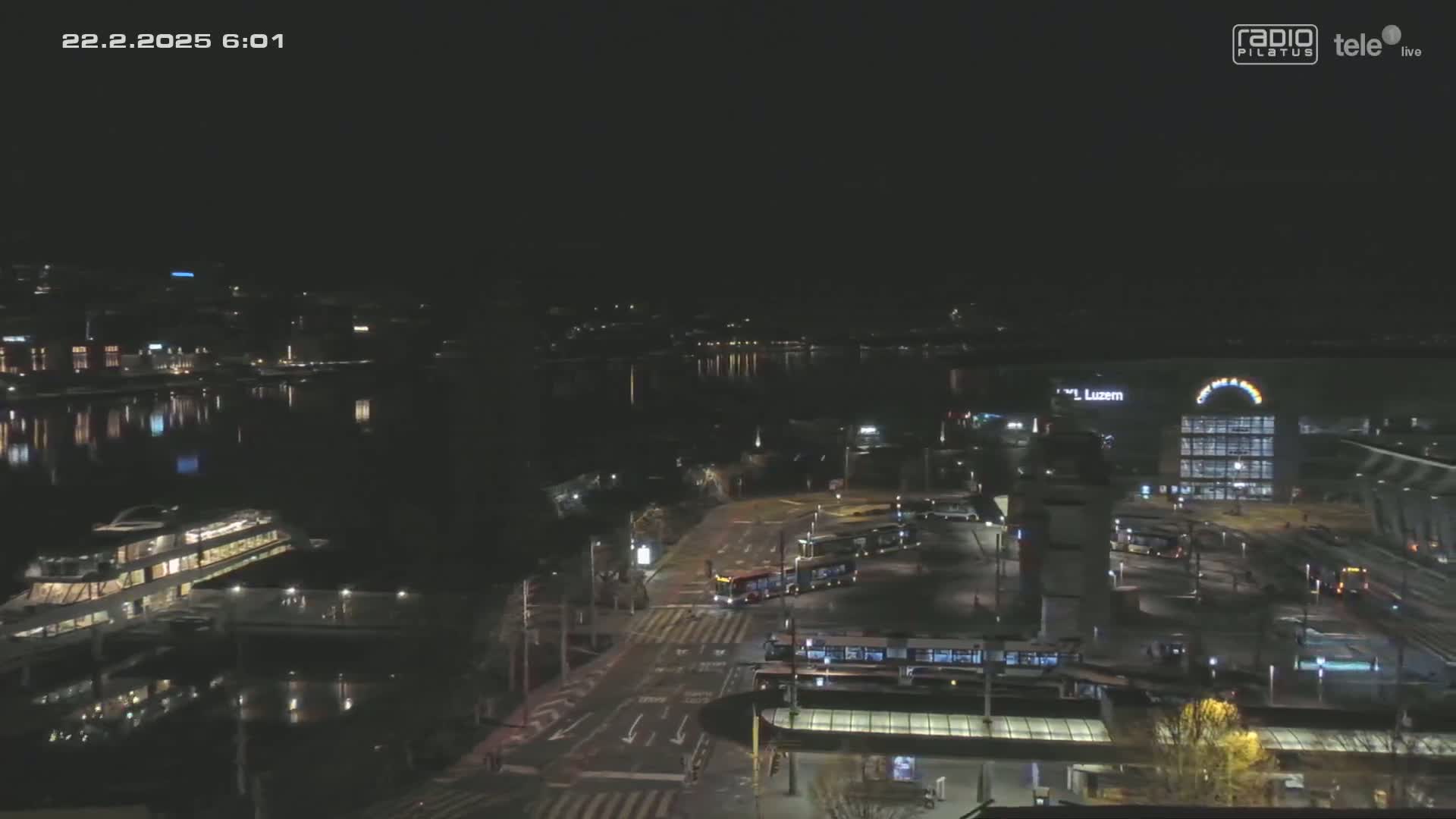 A nighttime aerial view shows a city waterfront with calm water reflecting lights, several trams at a station, and a large building illuminated.