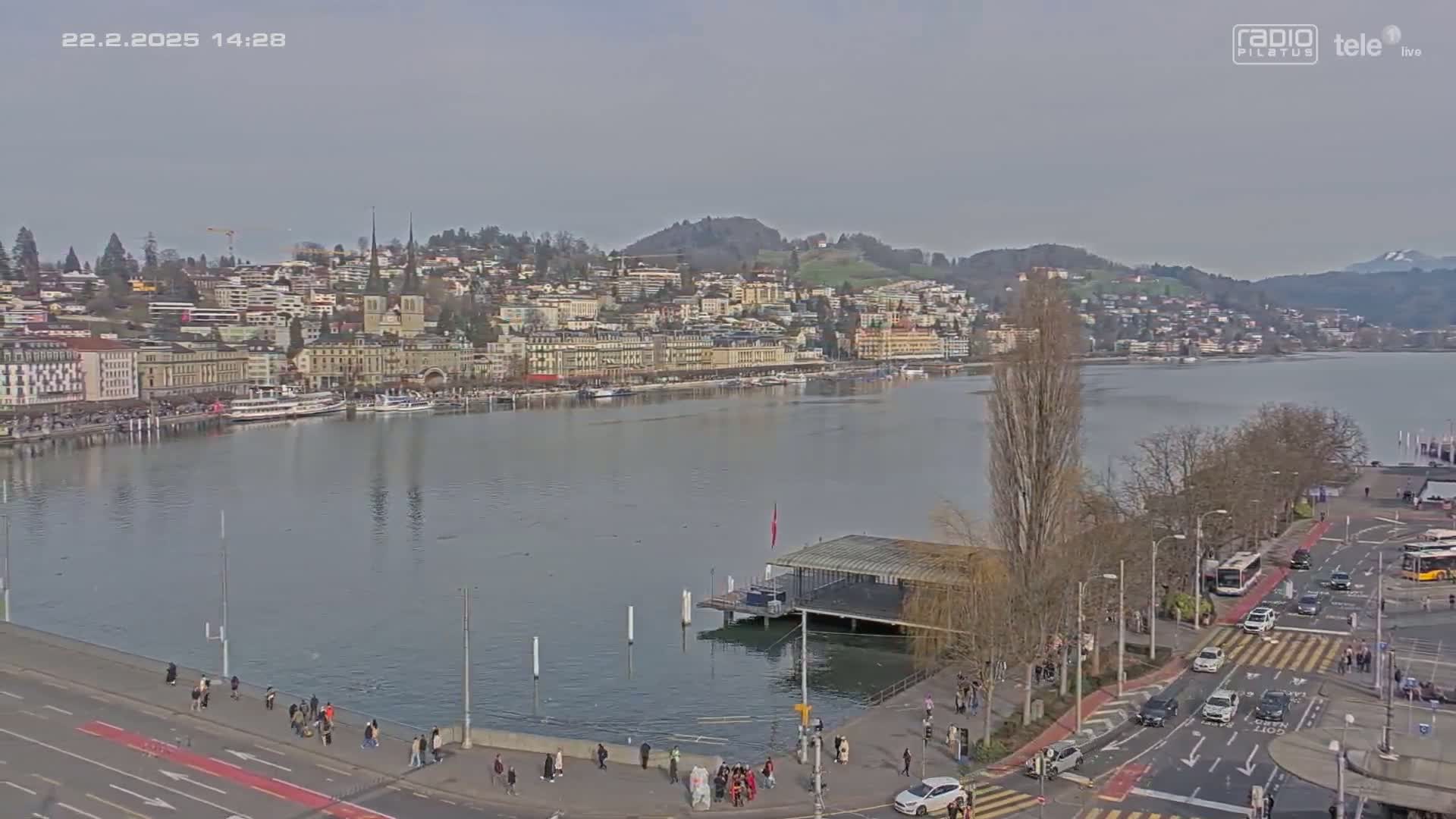 A waterfront city scene on a cloudy day shows a calm lake with boats, a town of buildings on a hillside, and a roadway with pedestrians and vehicles.
