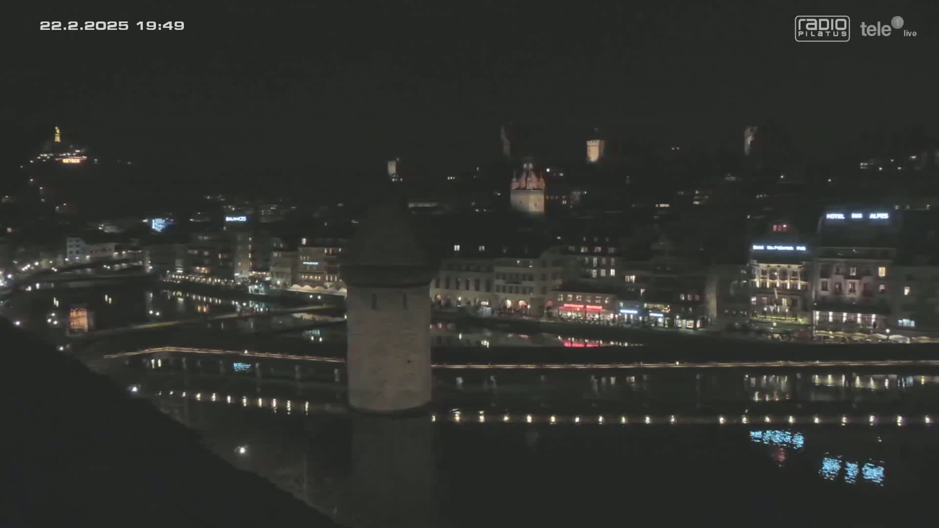 A nighttime, high-angle view of a city beside a body of water, showing the lights of buildings reflected in the dark water.
