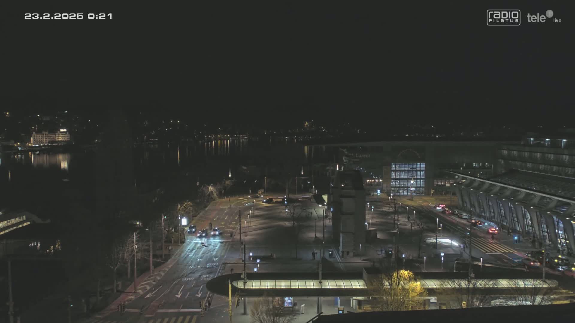 A nighttime, aerial view shows a mostly empty city square and roadway near a body of water, with a few vehicles visible and some buildings in the background under a clear, dark sky.