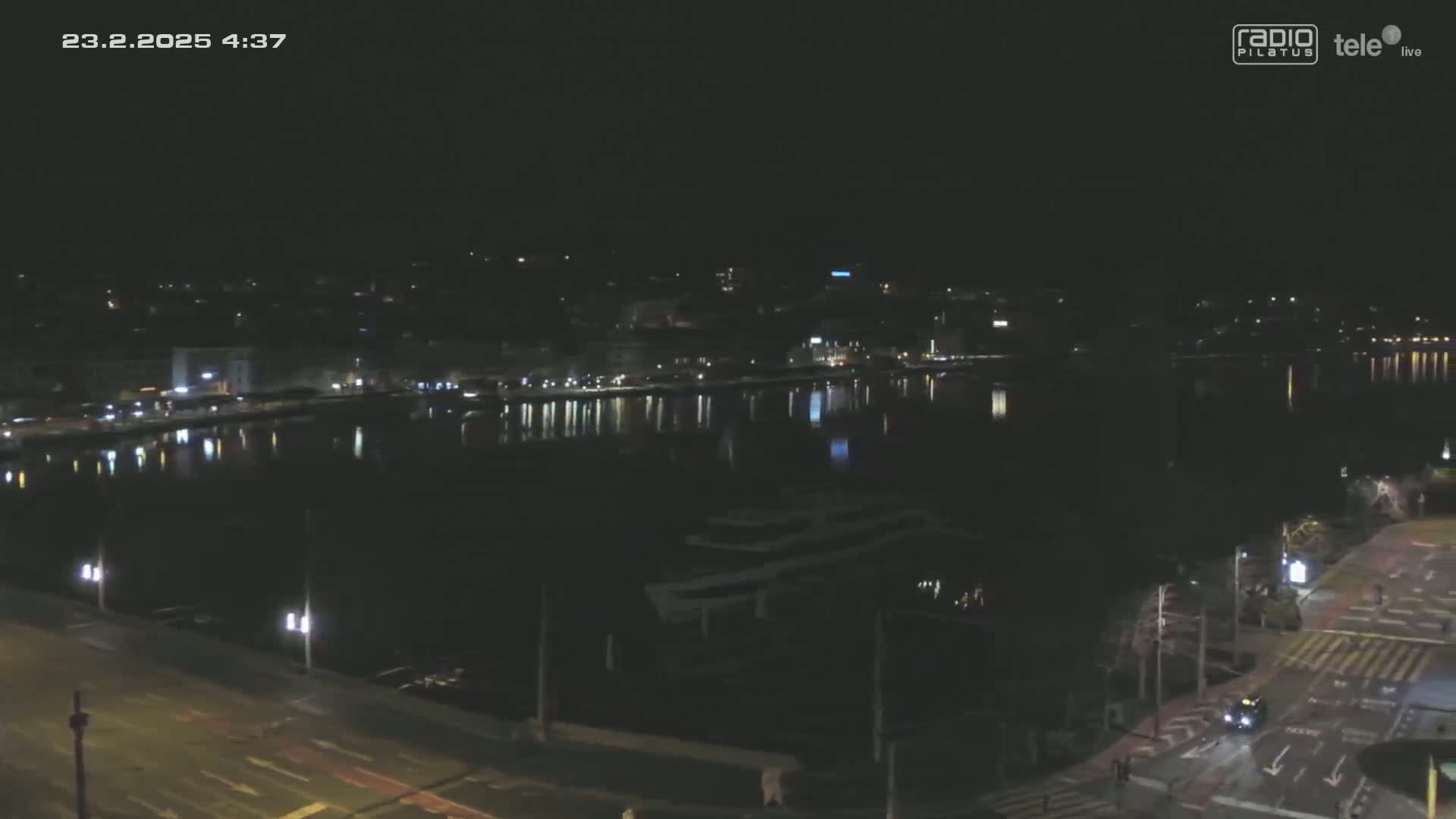 A nighttime, high-angle view shows a calm body of water reflecting the lights of a city across a roadway.