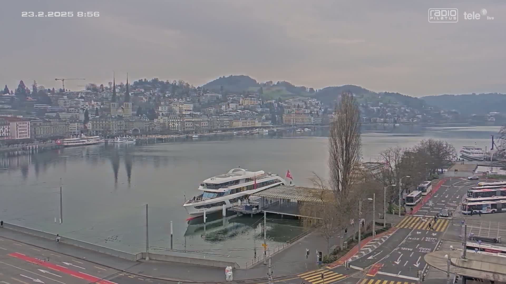 A large passenger boat is docked at a pier next to a calm lake, with a city and hills in the background under an overcast sky.