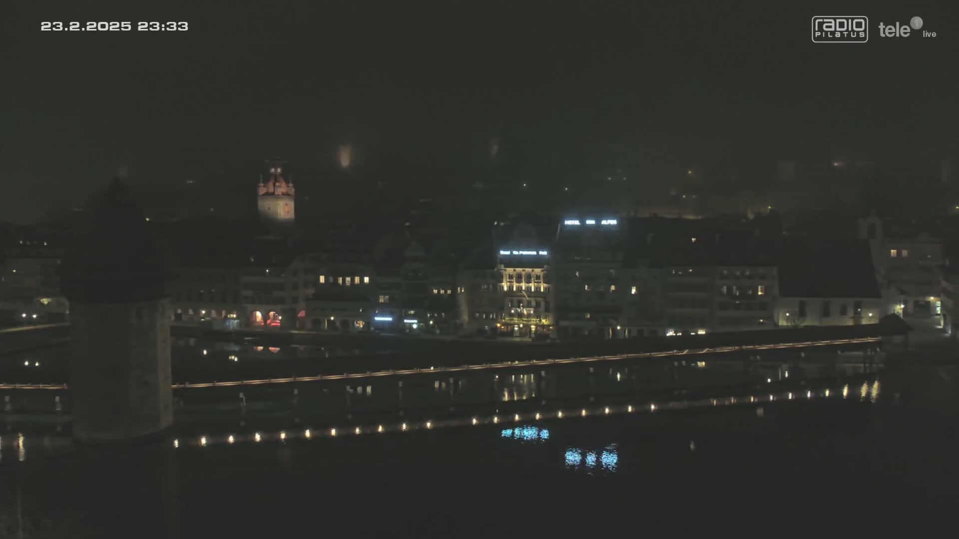 A nighttime, hazy view of a city along a body of water, featuring a bridge with lights, buildings, and a tower.