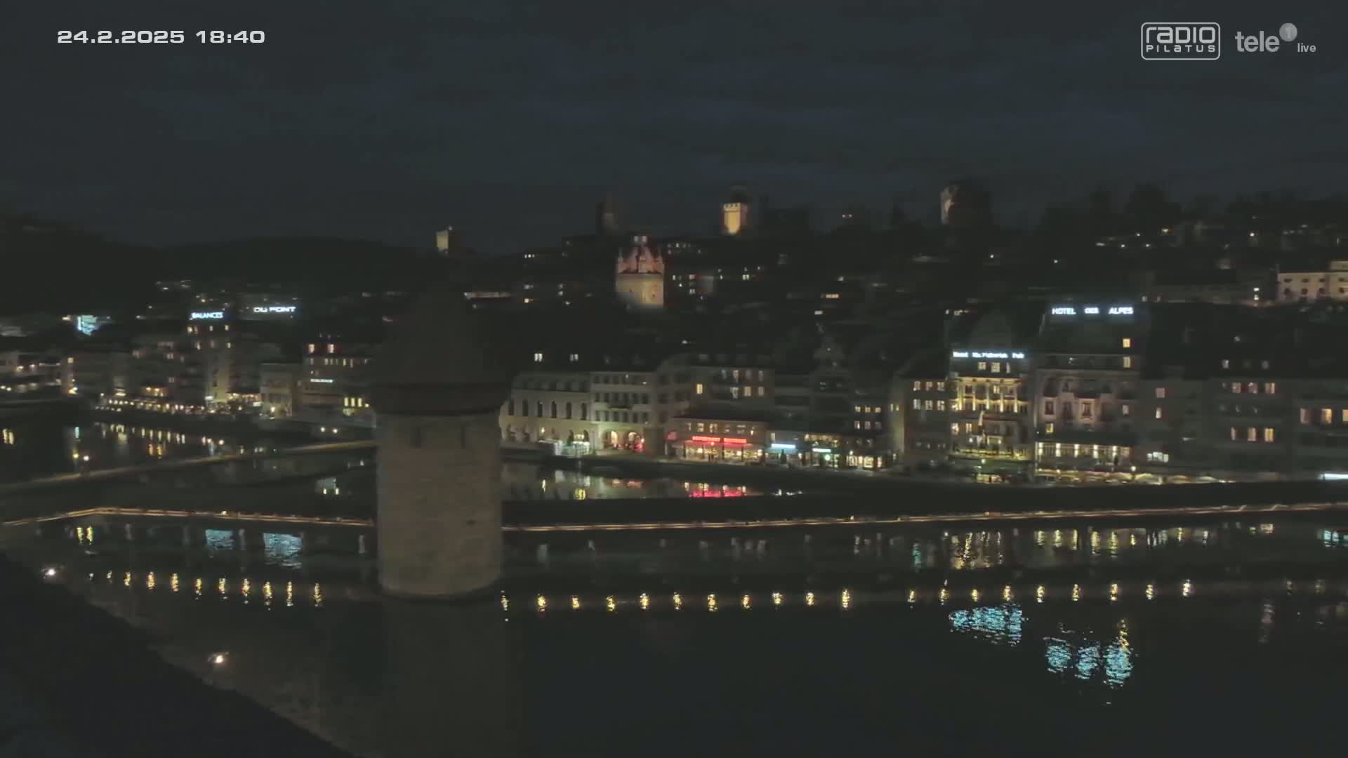 A nighttime view of a city illuminated along a calm river, reflecting lights on the water.