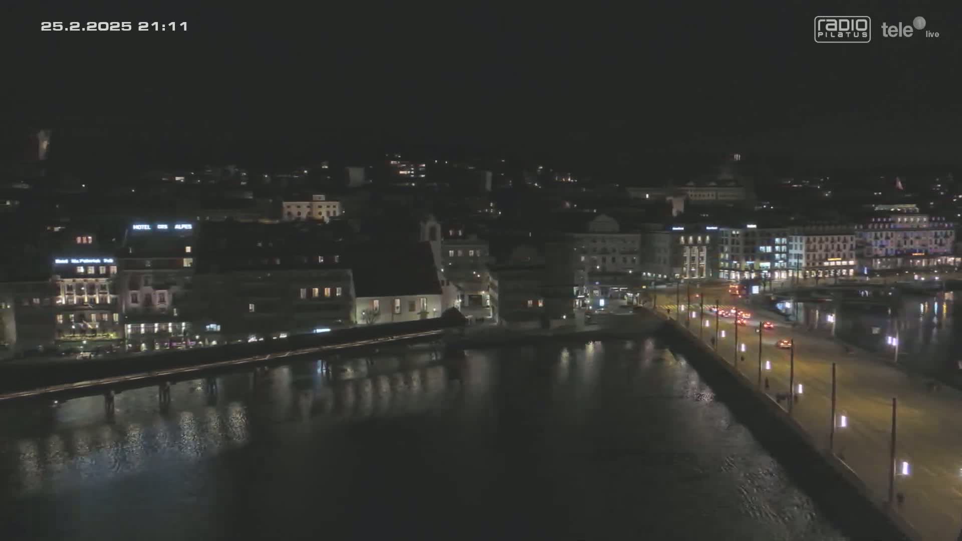 A nighttime aerial view shows a city waterfront with illuminated buildings and a bridge spanning a dark body of water under a clear sky.