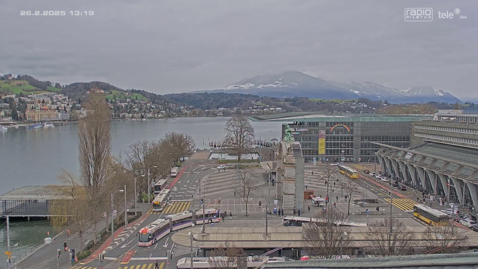 A city waterfront with buses and cars in a plaza, overlooked by buildings, under an overcast sky, with mountains in the background.