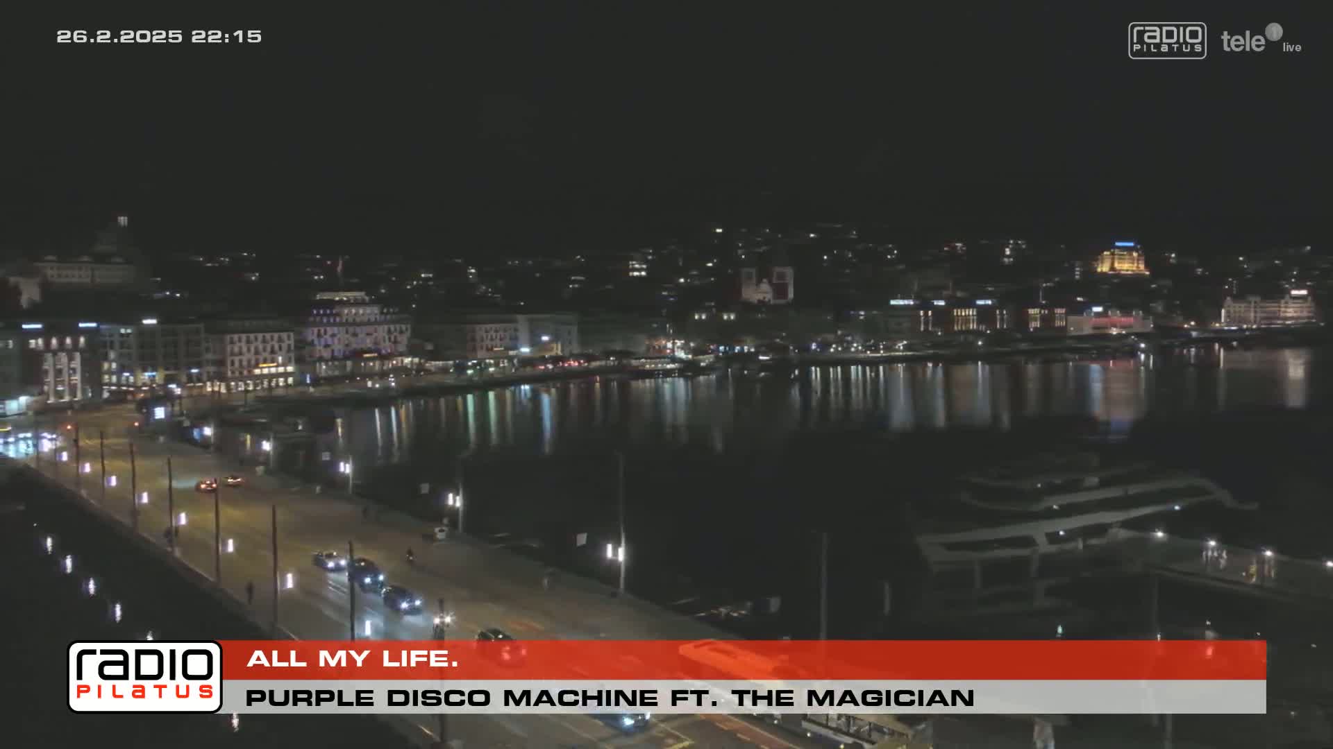 A nighttime view of a city waterfront with a calm body of water reflecting city lights, and a road with sparse traffic.