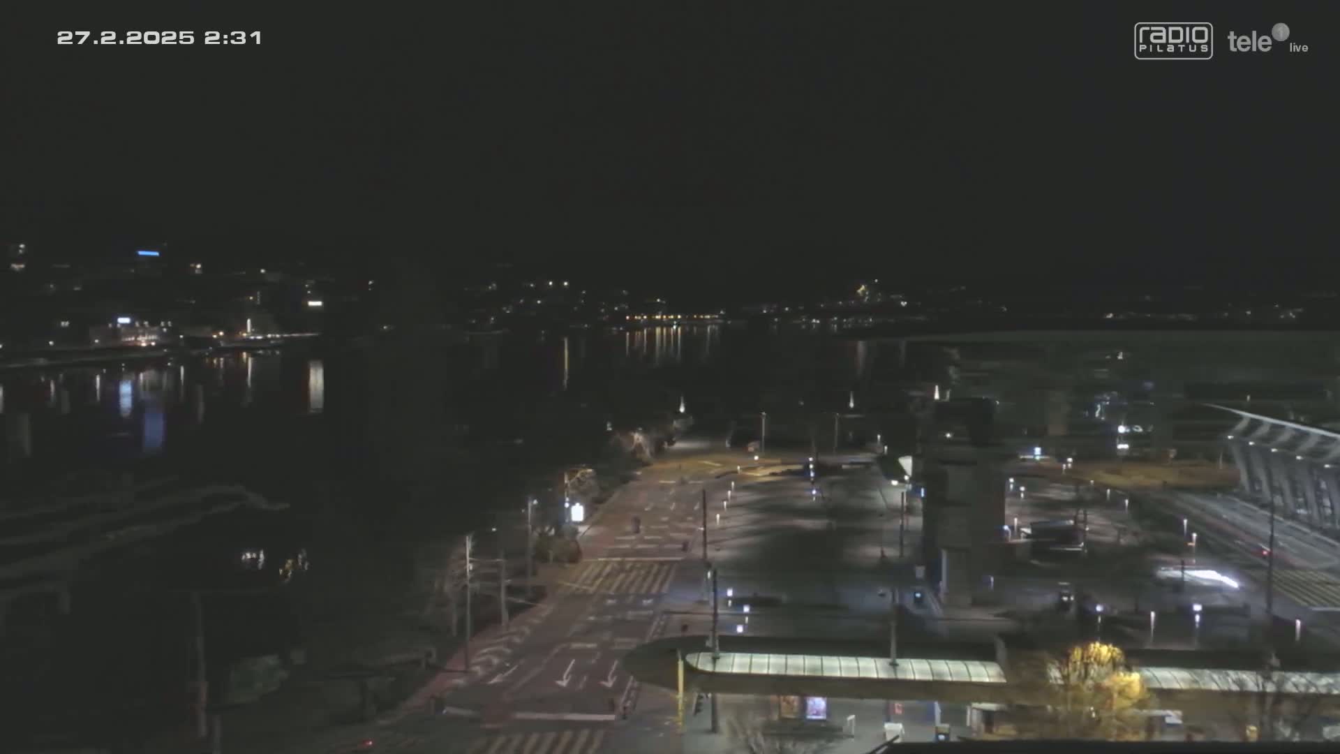A nighttime, high-angle view shows a mostly empty roadway and parking lot near a body of water with city lights reflecting on its surface.