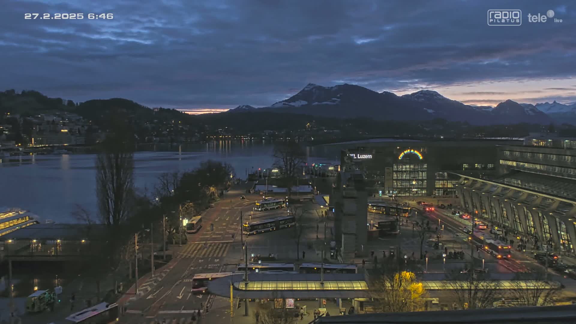 A twilight view of a city beside a lake, with mountains in the background under a partly cloudy sky.