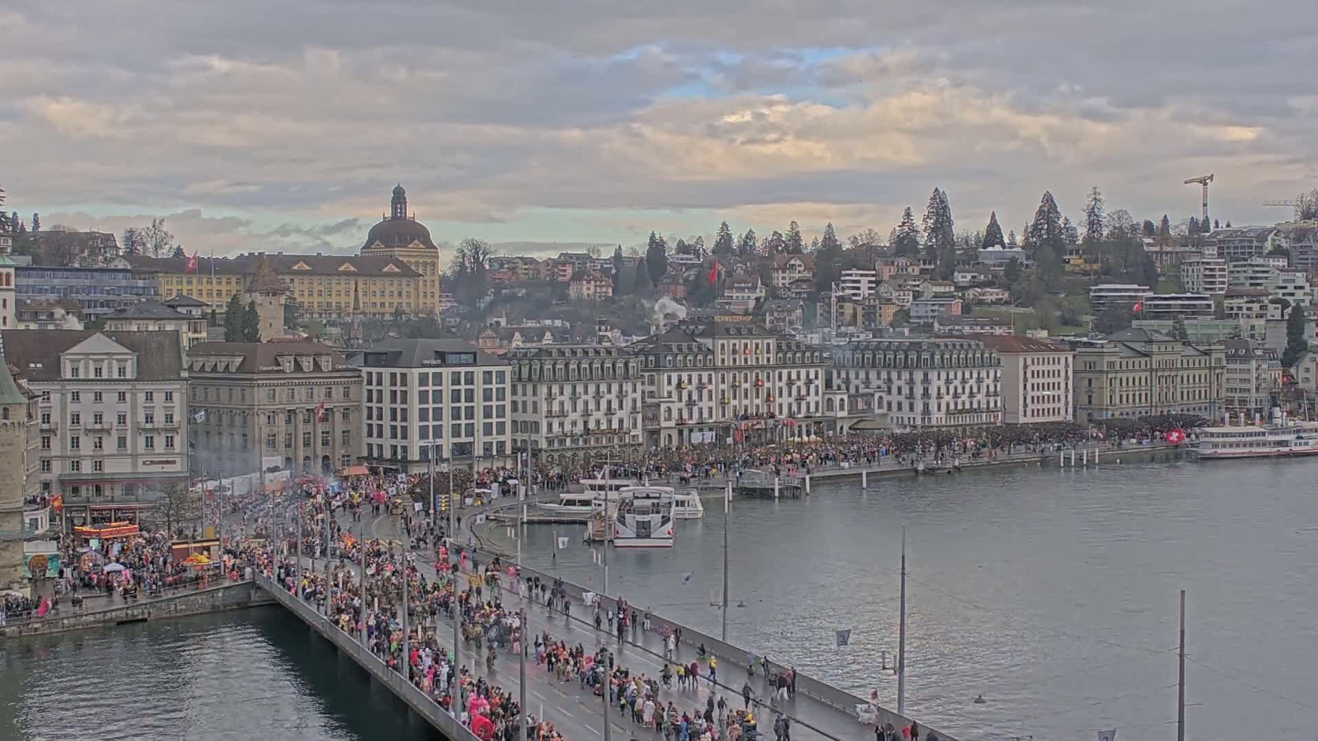 A large crowd of people is gathered on a bridge and along a waterfront, under a cloudy sky, in front of numerous buildings.