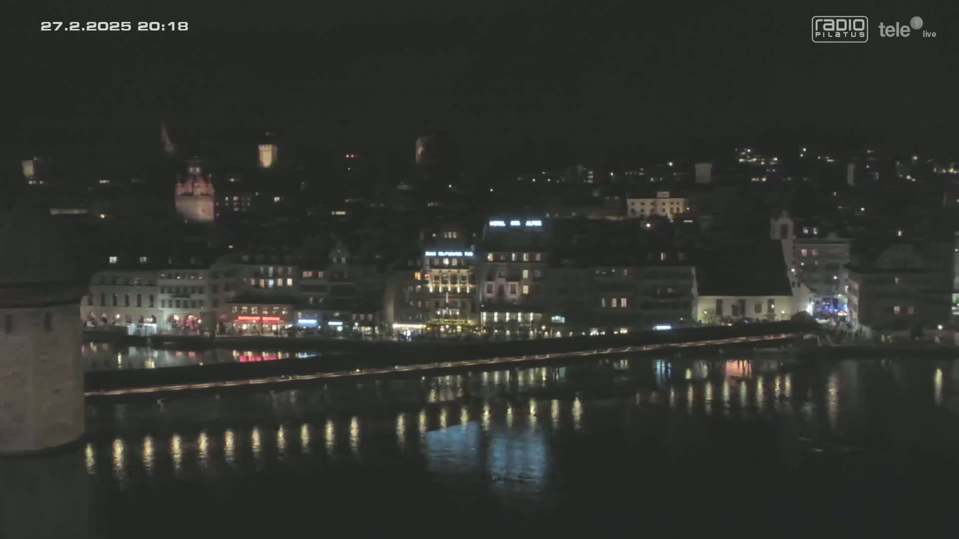A nighttime view of a city's waterfront, showing buildings with lights reflecting on the calm water.