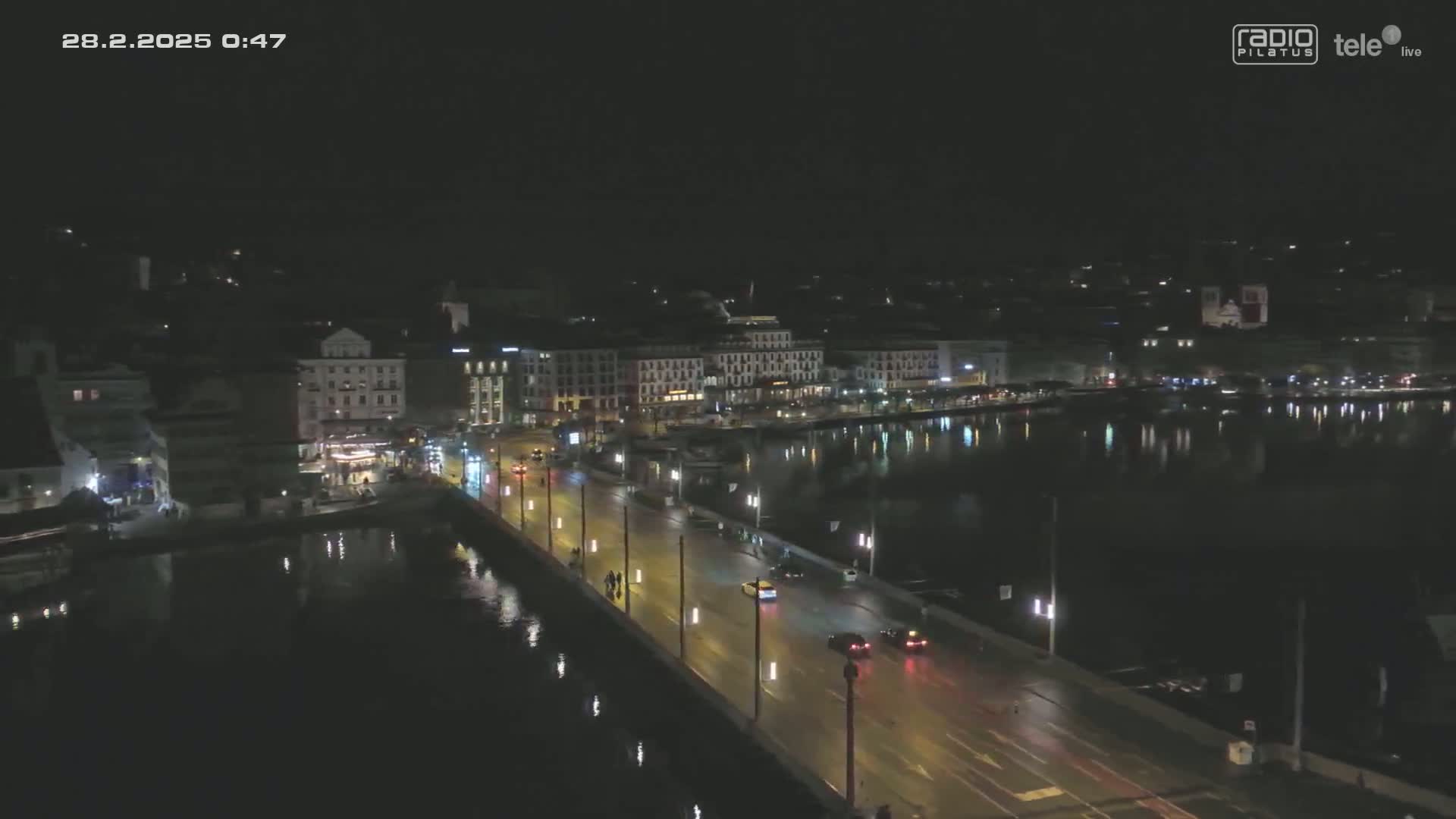 A nighttime, high-angle view of a city bridge with cars driving on it, beside a calm body of water reflecting city lights.