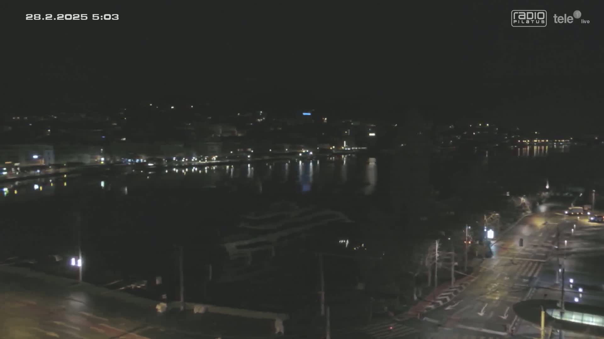 A nighttime view of a city waterfront, with dimly lit buildings and a dark body of water reflecting scattered lights.
