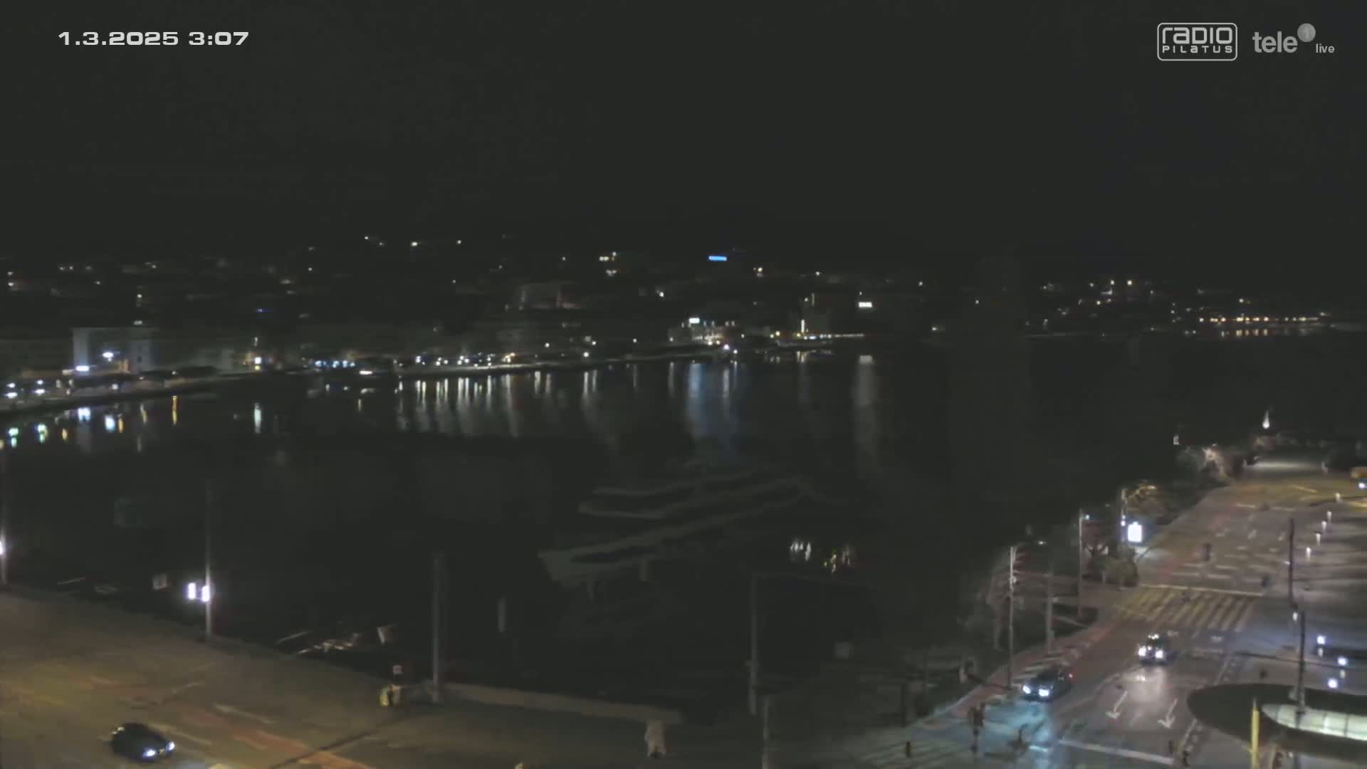 A nighttime view of a city waterfront, with calm water reflecting city lights under a clear, dark sky.