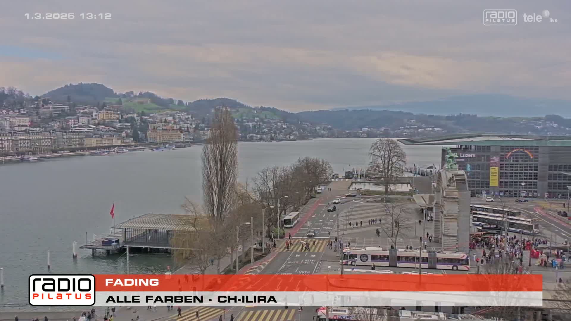 A city waterfront on an overcast day, showing a body of water, buildings, and a street scene with pedestrians and vehicles.