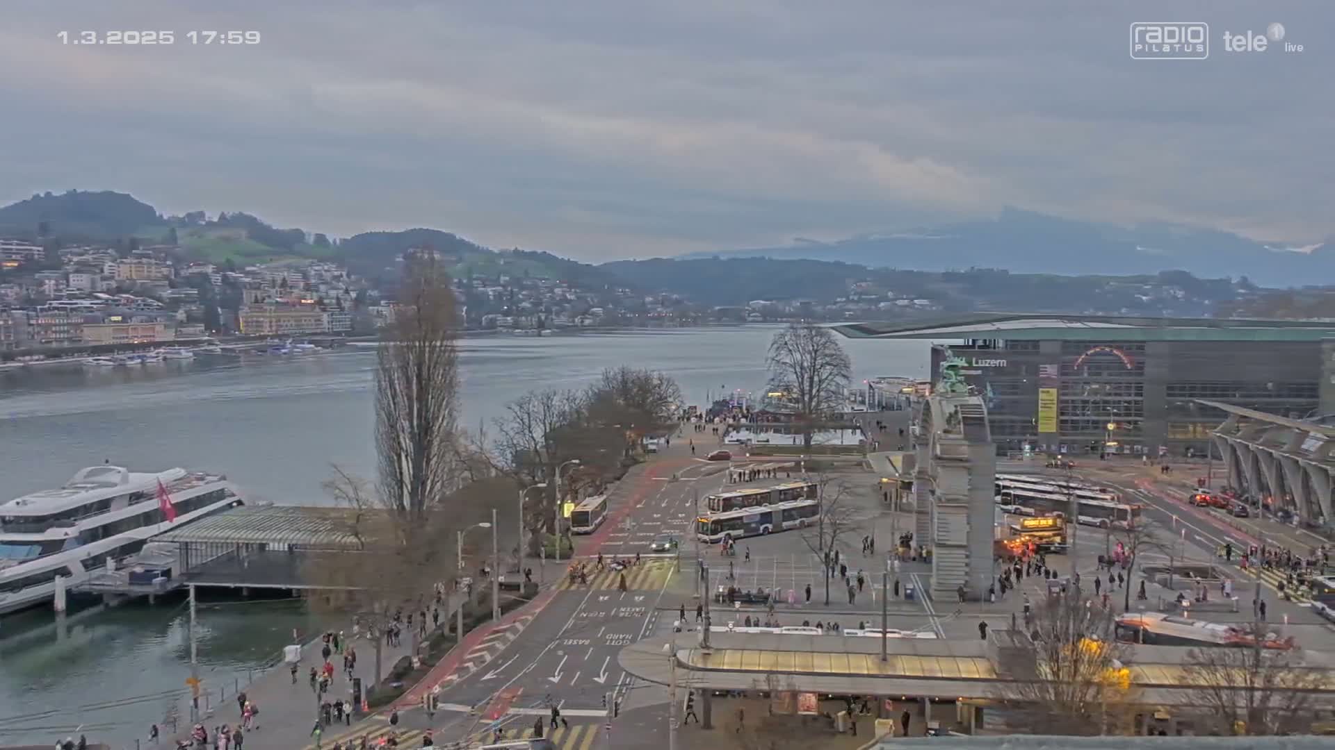 An overcast day reveals a lakeside city with a large body of water, numerous buses and pedestrians near a waterfront building, and a large passenger ship docked nearby.