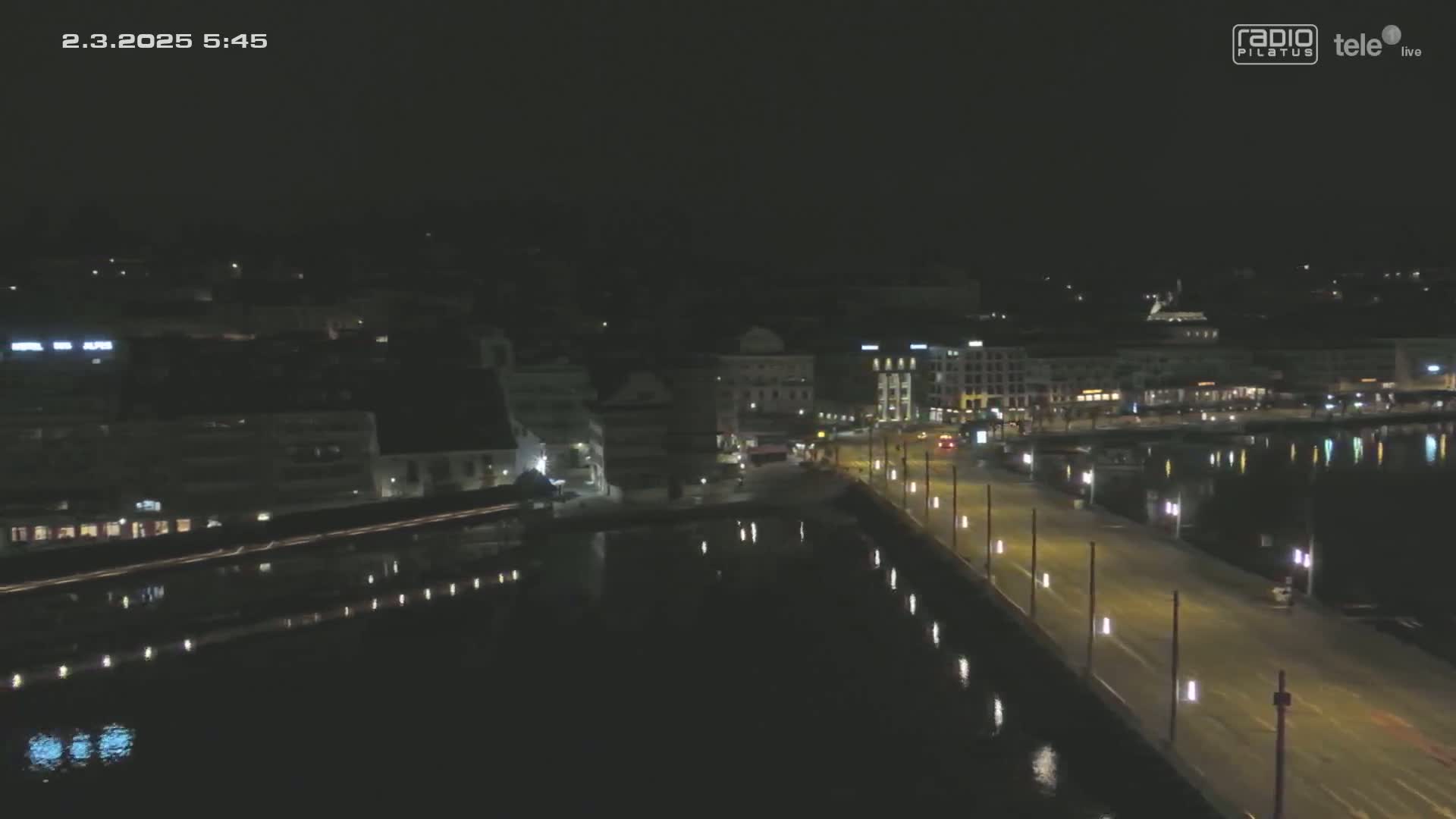 A nighttime aerial view shows a city waterfront with a long, illuminated roadway next to calm, dark water reflecting the city lights.