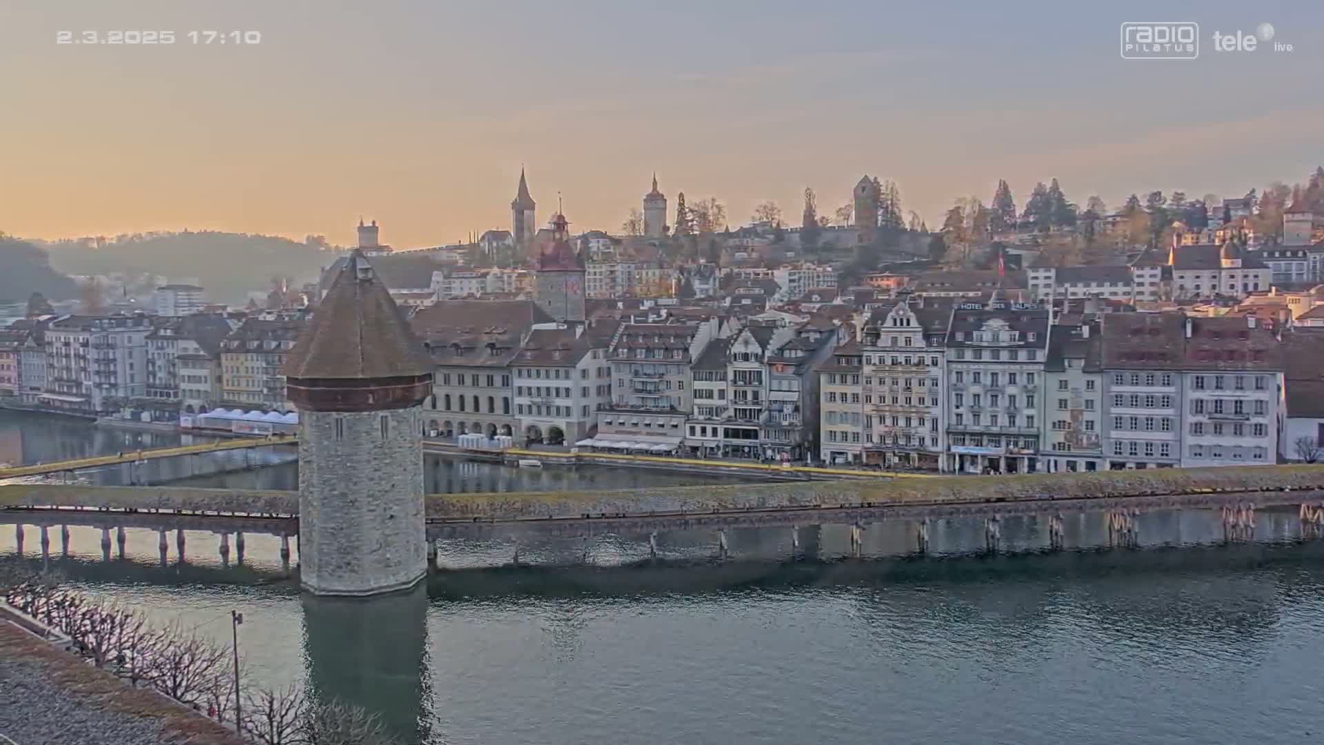 A stone tower at the end of a bridge sits on a calm river in front of a city with many buildings under a soft, dusky sky.