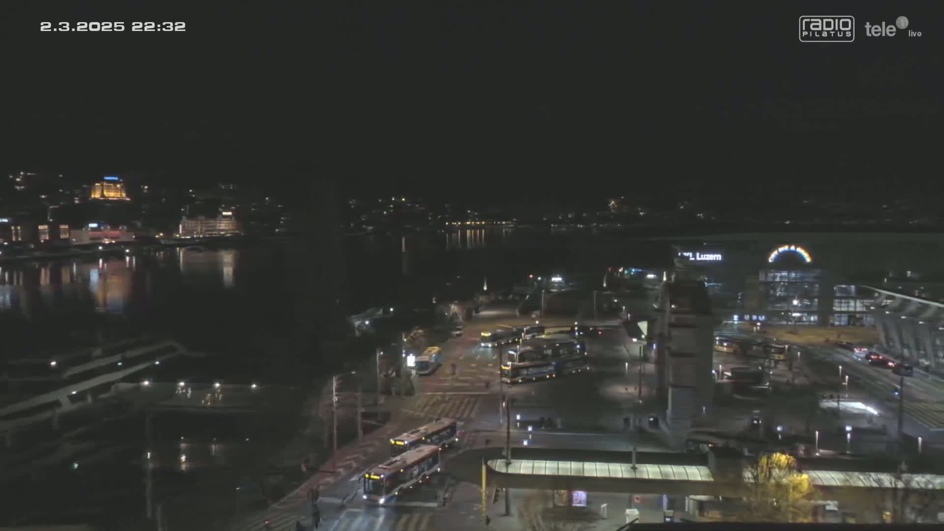 A nighttime view of a city’s waterfront, showing a body of water reflecting lights from buildings and vehicles on the streets near a transportation hub.