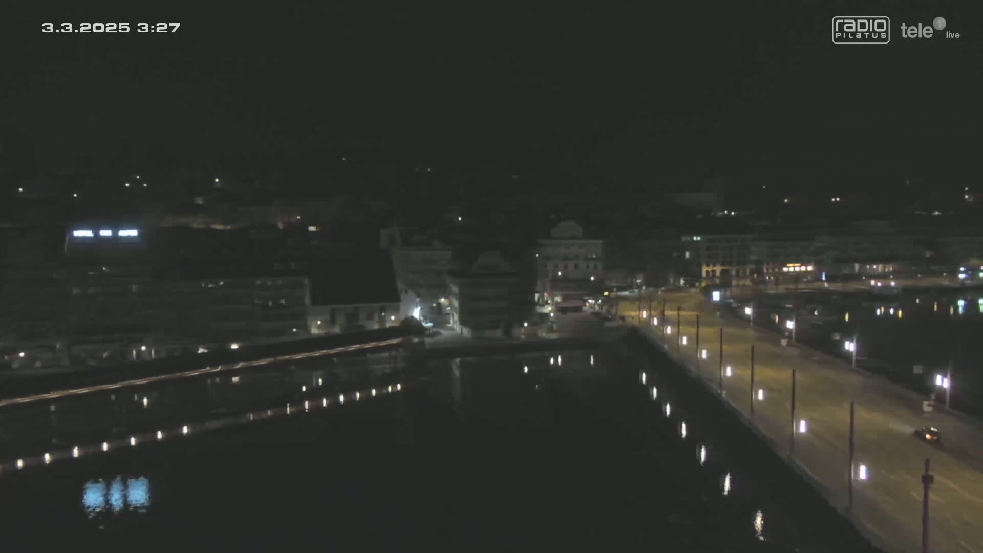 A nighttime, high-angle view shows a city's waterfront at night, with calm water reflecting the lights of buildings and a roadway.