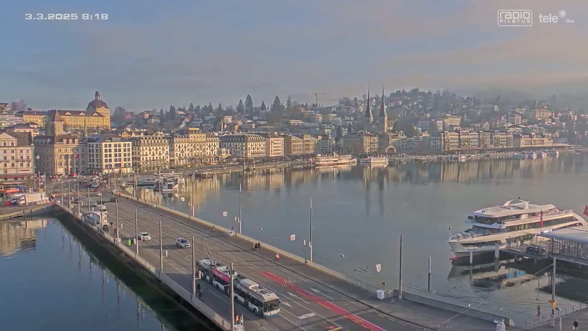 A long bridge spans a calm body of water, with a city of multi-story buildings and several boats in the background under a clear, slightly hazy sky.