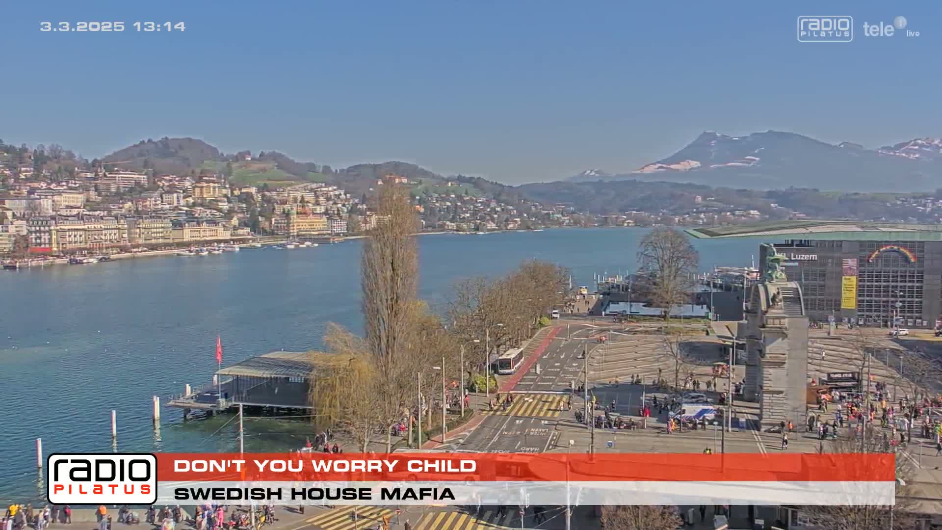 A sunny day overlooks a city by a lake with mountains in the background, and a large plaza with many people is visible in the foreground.