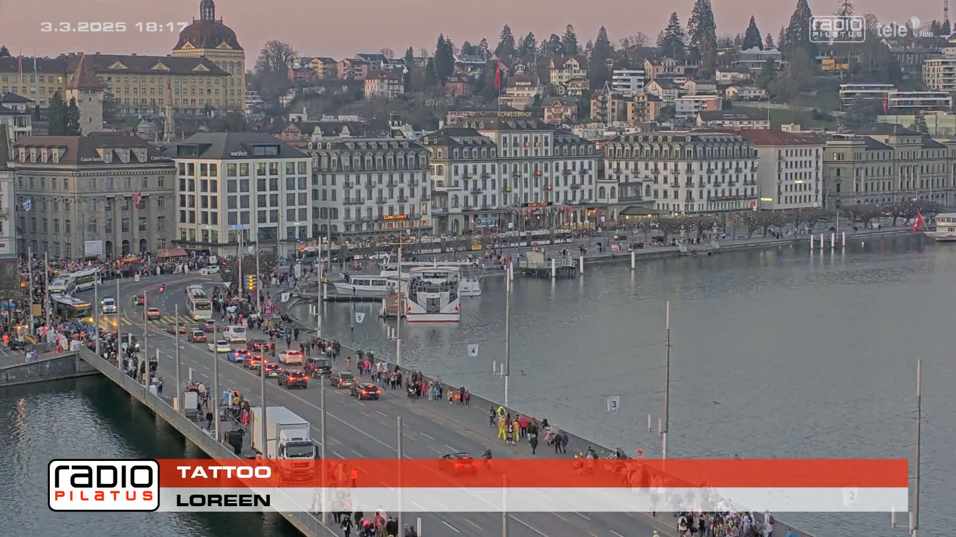 A large crowd of people walks along a bridge and a waterfront road next to a calm lake at dusk, with numerous buildings and boats visible in the background.