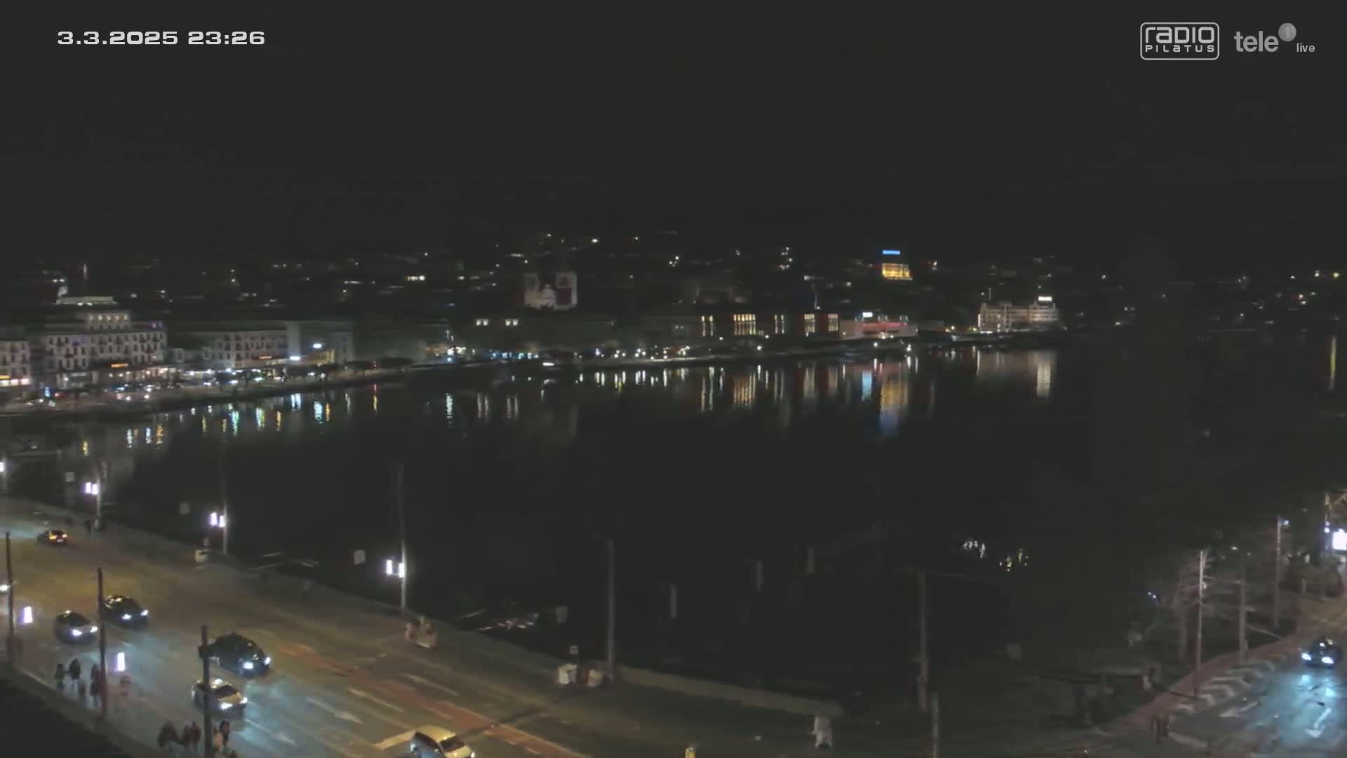 A nighttime view of a city waterfront, with calm water reflecting the lights of buildings along a roadway.