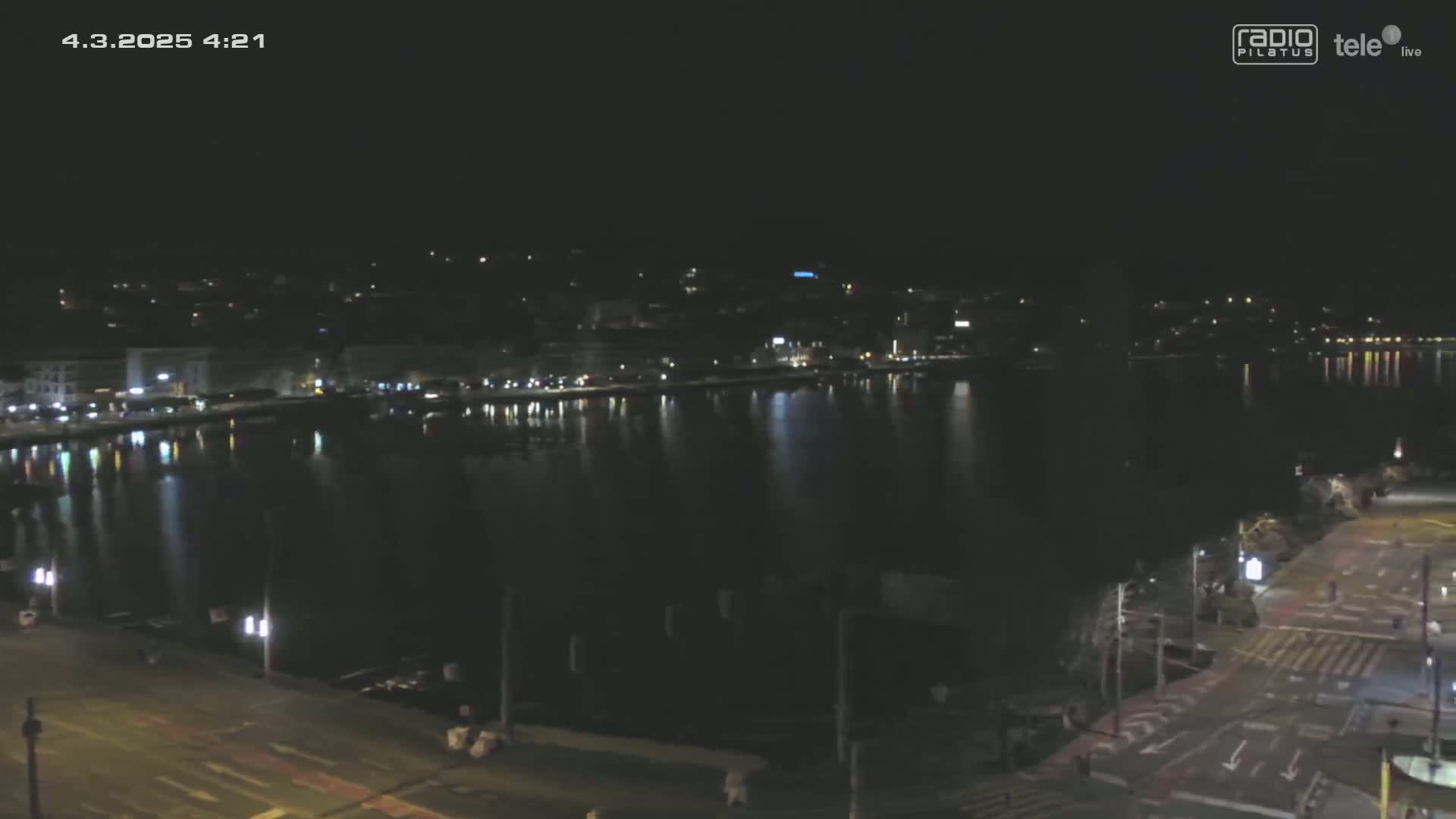 A nighttime view of a calm body of water reflecting city lights, with a road and buildings visible in the foreground.
