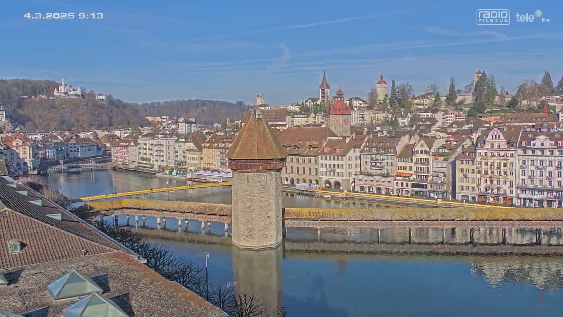 A stone tower stands on a covered bridge spanning a calm river, reflecting the cityscape and a clear blue sky beyond it.